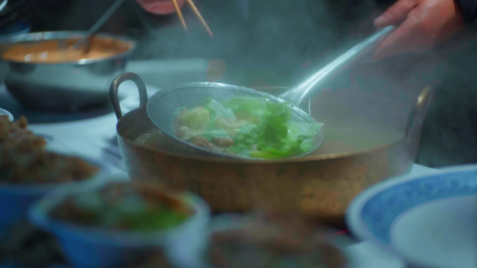 Close-up of a steaming pot over a stove with fresh green vegetables inside, surrounded by small bowls of various dishes.