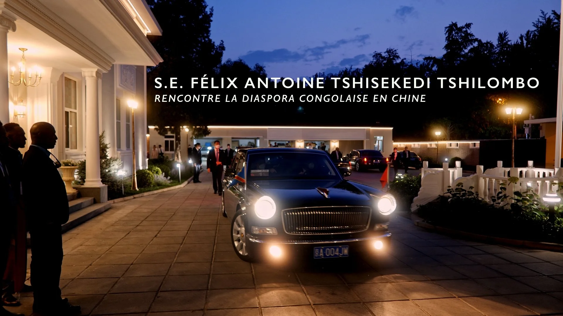 Exterior scene at night with a black luxury car in front of a white building, string lights, and several people standing around, some in suits, during a formal event.