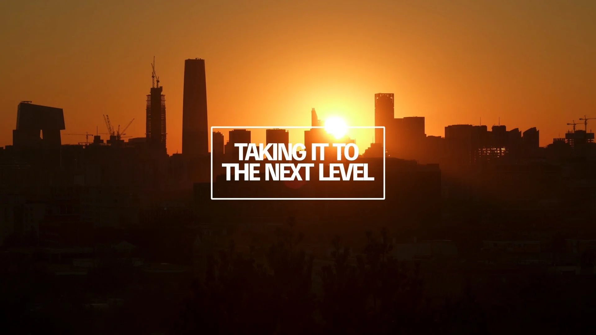 Sunset over a city skyline with tall buildings and construction cranes, with the text 'Taking it to the next level' displayed in the foreground.