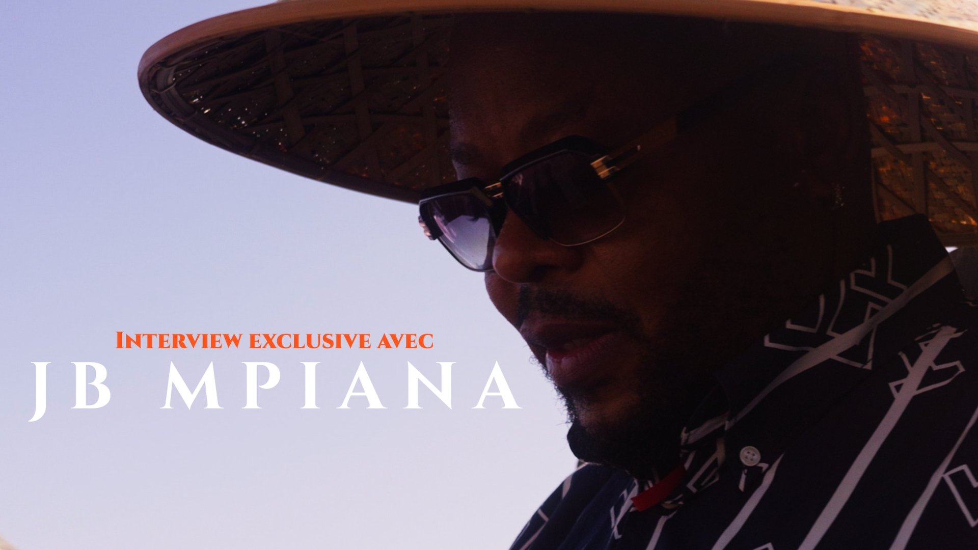 Close-up of a man wearing sunglasses and a patterned shirt, with text overlaying that reads 'Interview exclusive avec JB M PIANA'.