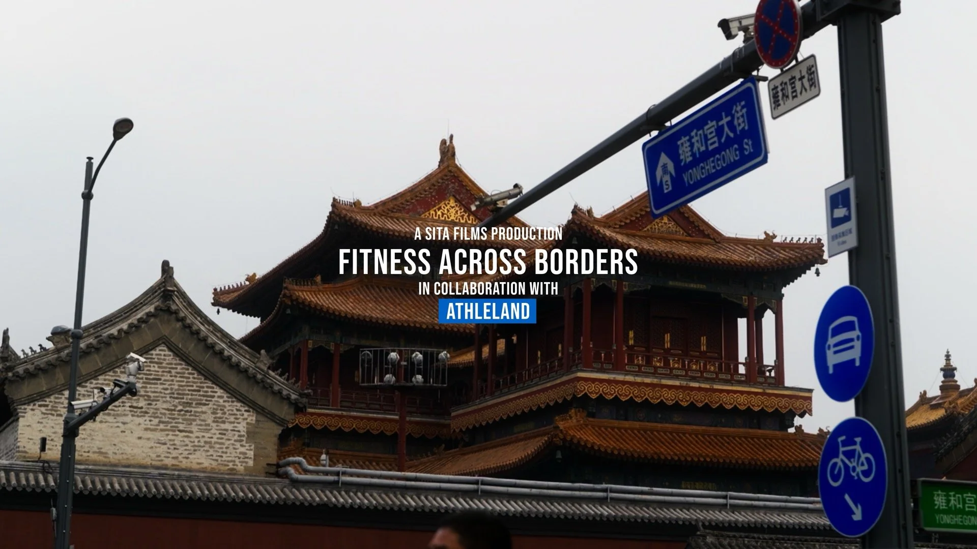 Title card for "Fitness Across Borders" film with a background of traditional Chinese architecture, street signs in Chinese and English, and overcast sky.