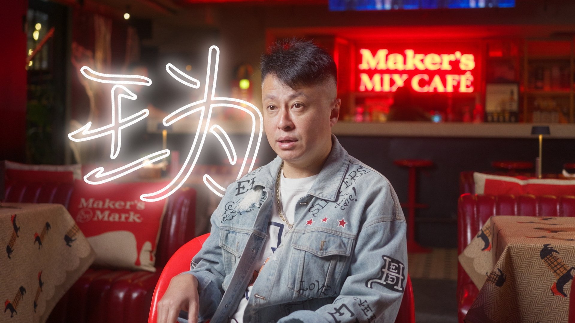 A man sitting in a red chair inside a cafe named Maker's Mix Cafe. He is wearing a denim jacket with various patches and designs. Behind him, there is a neon sign with Chinese characters and a red sign that reads Maker's Mix Cafe. The cafe has red ch