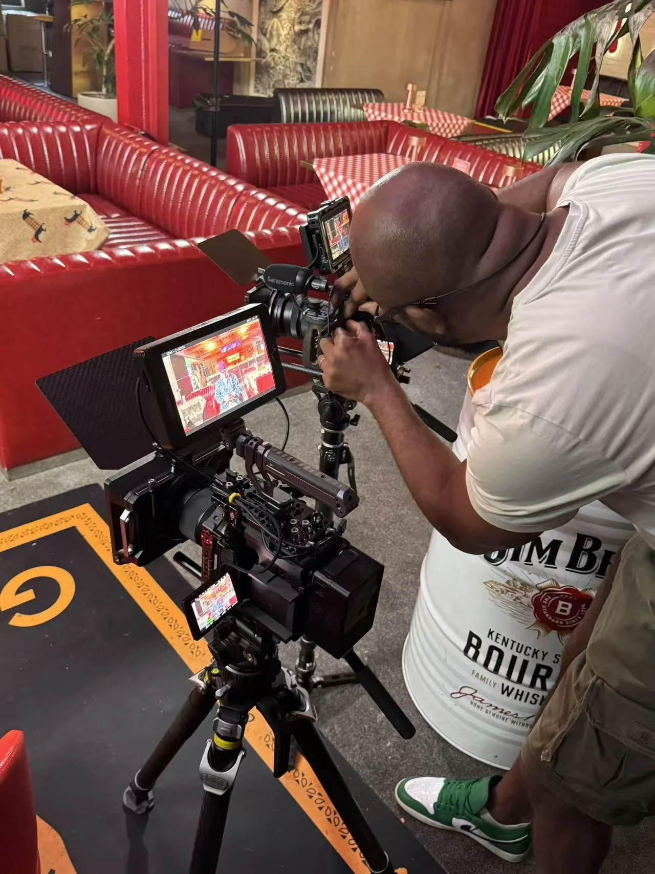 A person using a professional camera mounted on a tripod to shoot a scene inside a restaurant with red booth seating and checkered tablecloths.