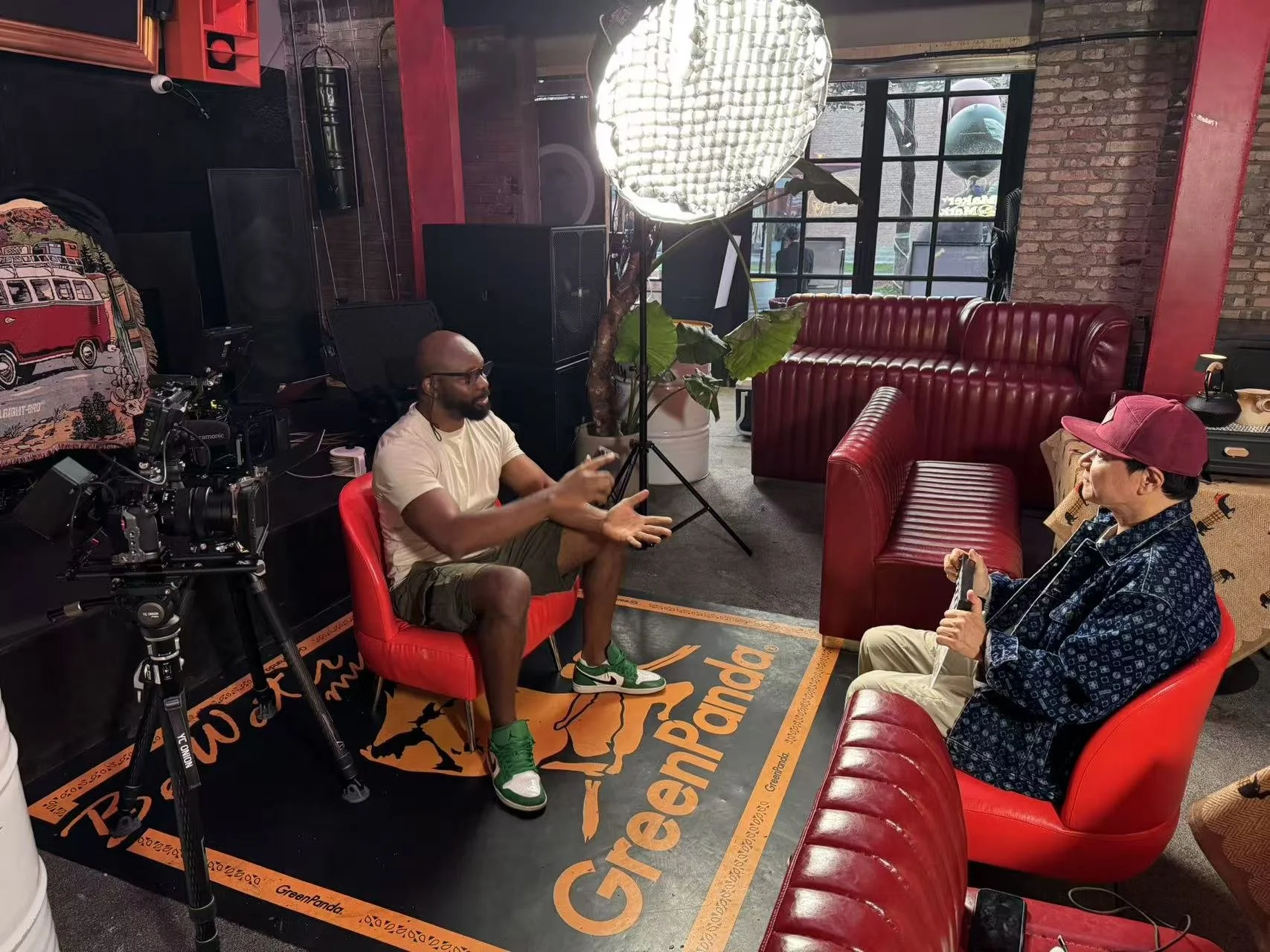 Two men are sitting and talking in a cozy lounge or studio with brick walls. One man in a beige shirt and shorts is gesturing with his hands, while the other in a patterned shirt, beige pants, and a maroon cap is holding a phone. A camera on a tripod is pointed at them, and there is professional lighting. There are red leather couches, a large potted plant, and large windows showing an urban exterior.