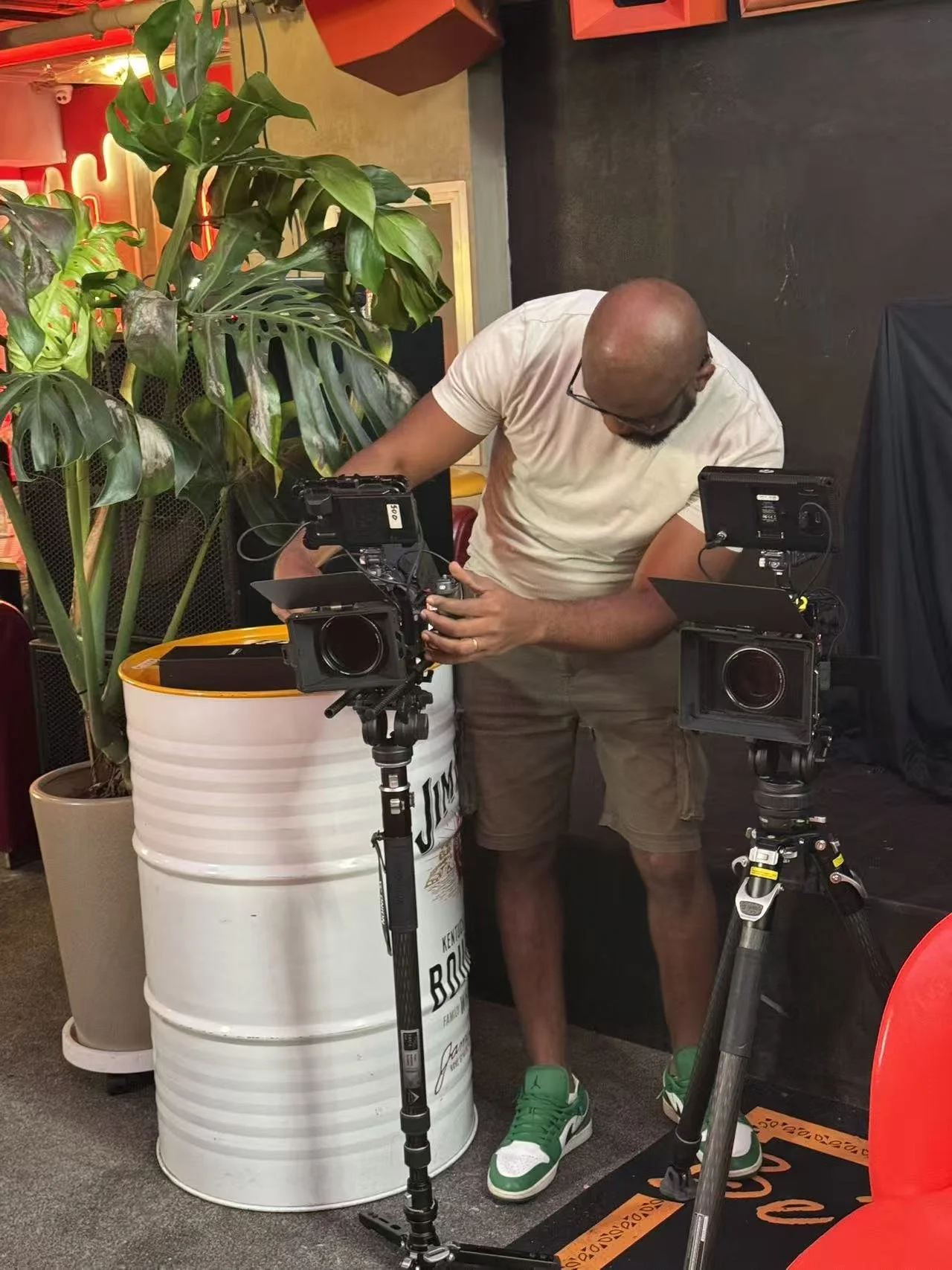 A man adjusting camera equipment in an indoor space with plants and colorful decor.