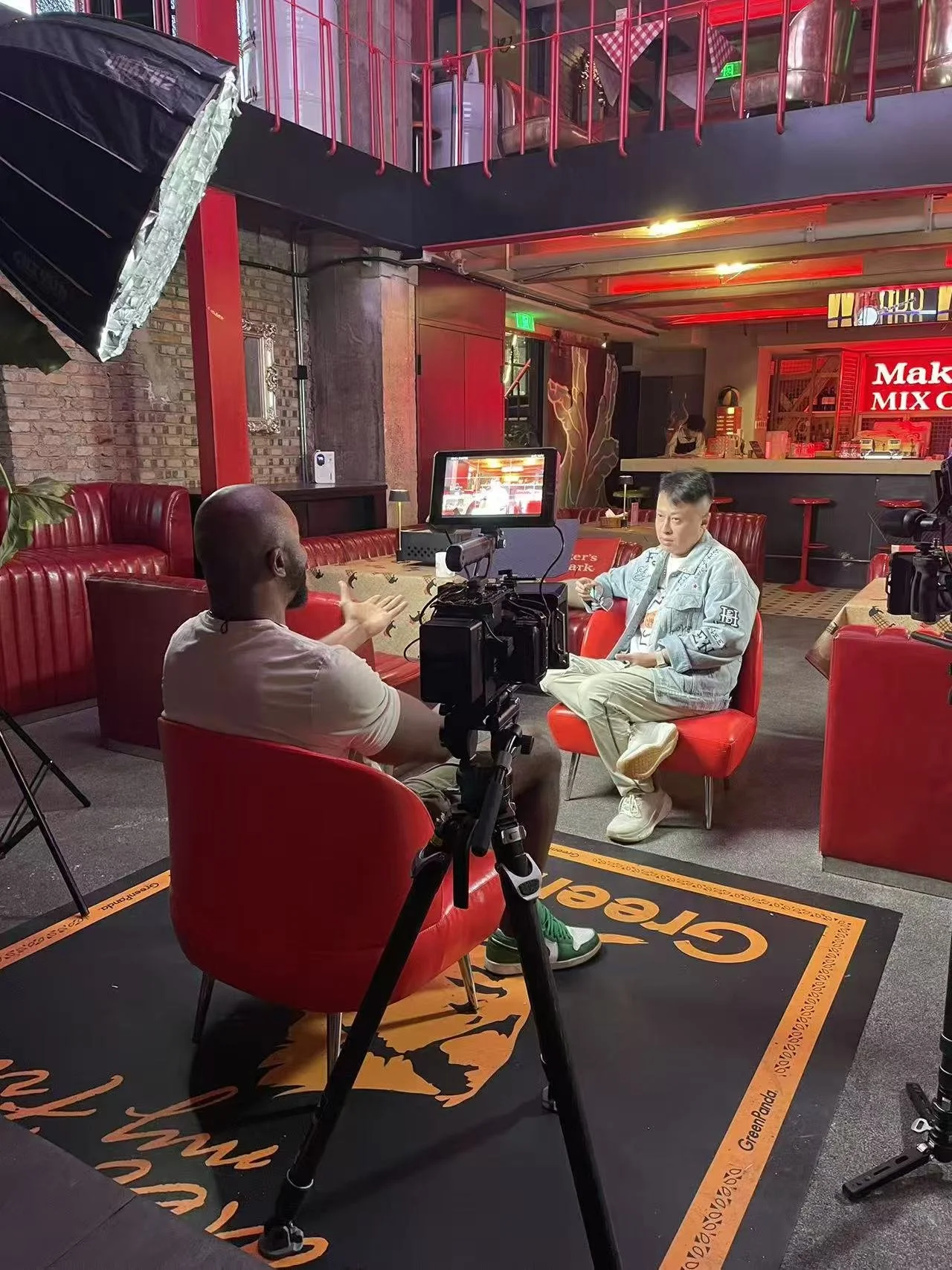 Two men are seated in a red booth set up for an interview or filming in a dimly lit, modern indoor space with red neon lighting. One man is talking, using hand gestures, while the other man is listening and holding a phone. A camera, lighting, and monitor are positioned to record the scene. The background features a bar area with a red neon sign and decorative artwork on the walls.