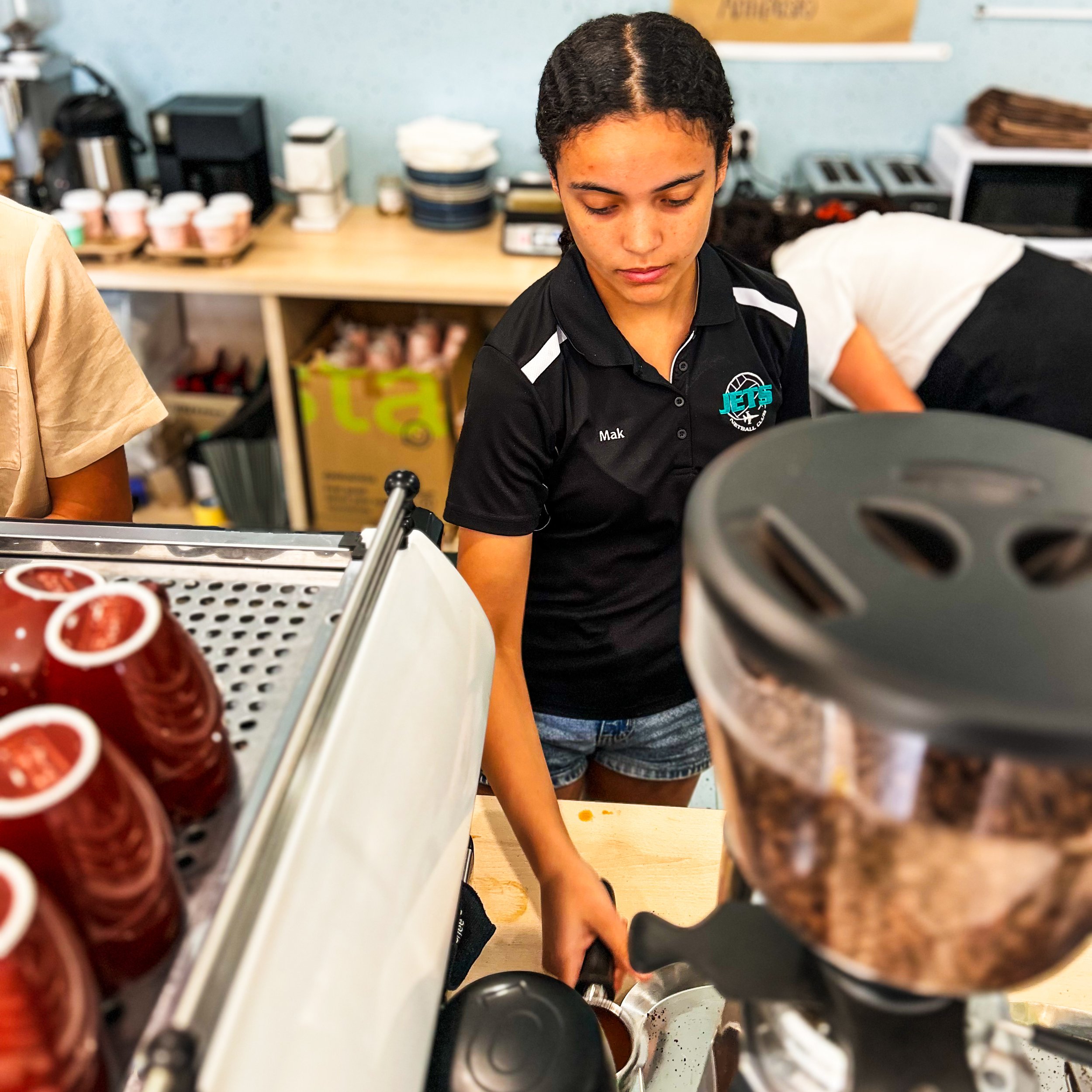 A young woman with dark curly hair, wearing a black jersey with a logo and the name 'Mak', looking down while working with kitchen utensils in a kitchen or cafe setting. There are shelves with cups and containers and kitchen appliances in the background.