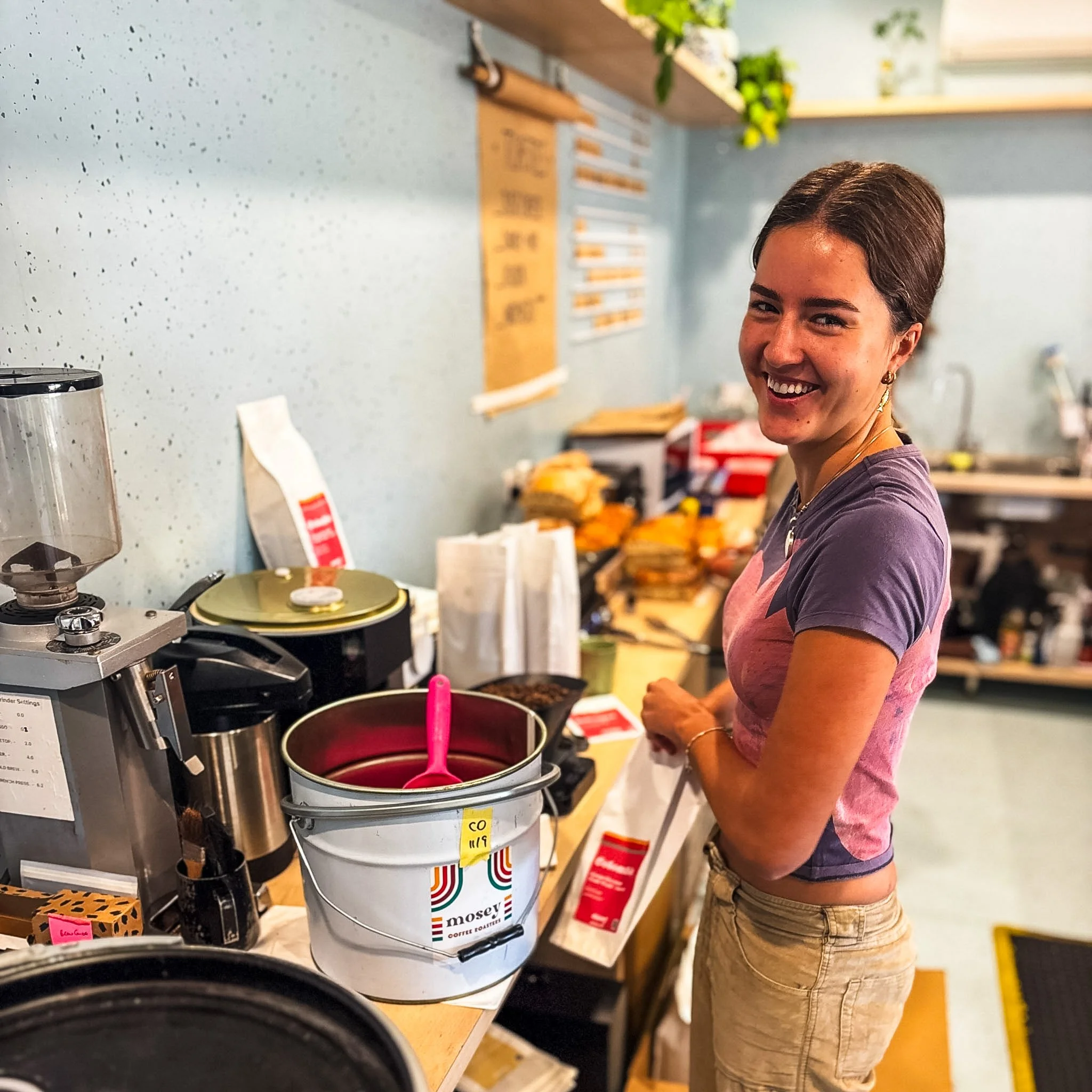 Woman smiling and working at a coffee counter in a cafe or coffee shop, surrounded by coffee-related equipment and products.