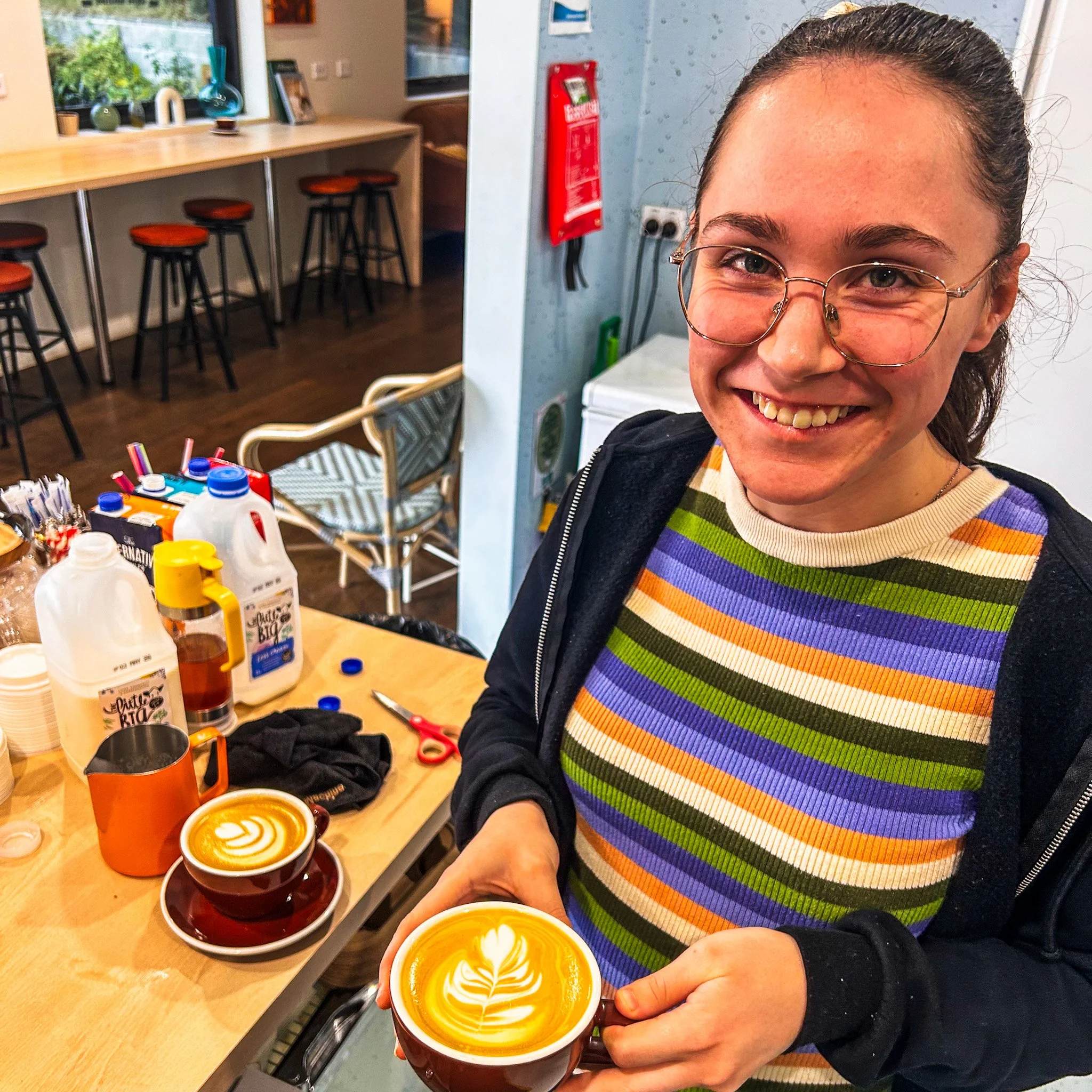 Woman with glasses holding a coffee cup with latte art in a café. The counter behind her has bottles of milk and other items, with stools along the counter and a window with plants outside.