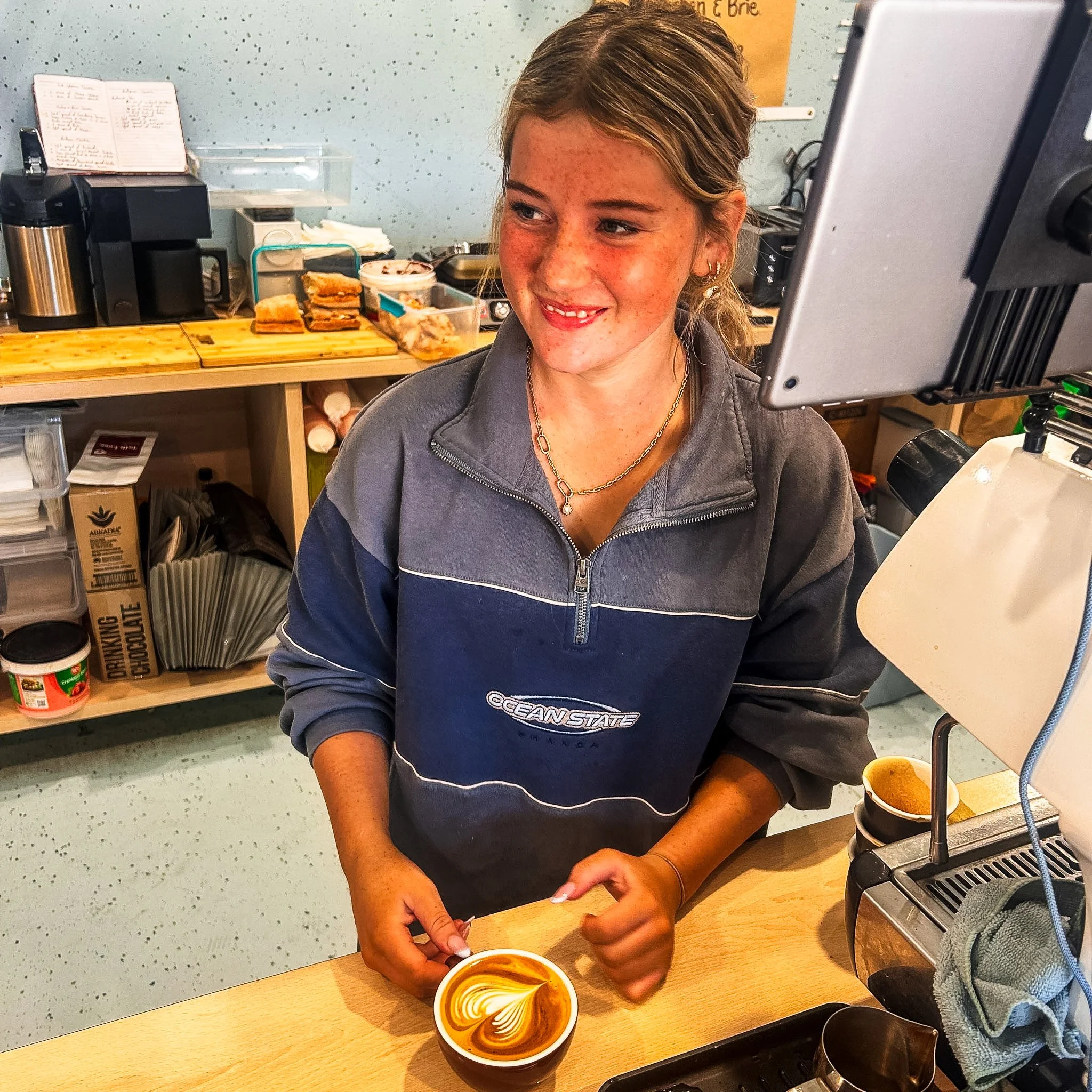 A young woman with light brown hair and freckles, smiling, making art on a coffee with foam art, at a coffee shop or cafe.