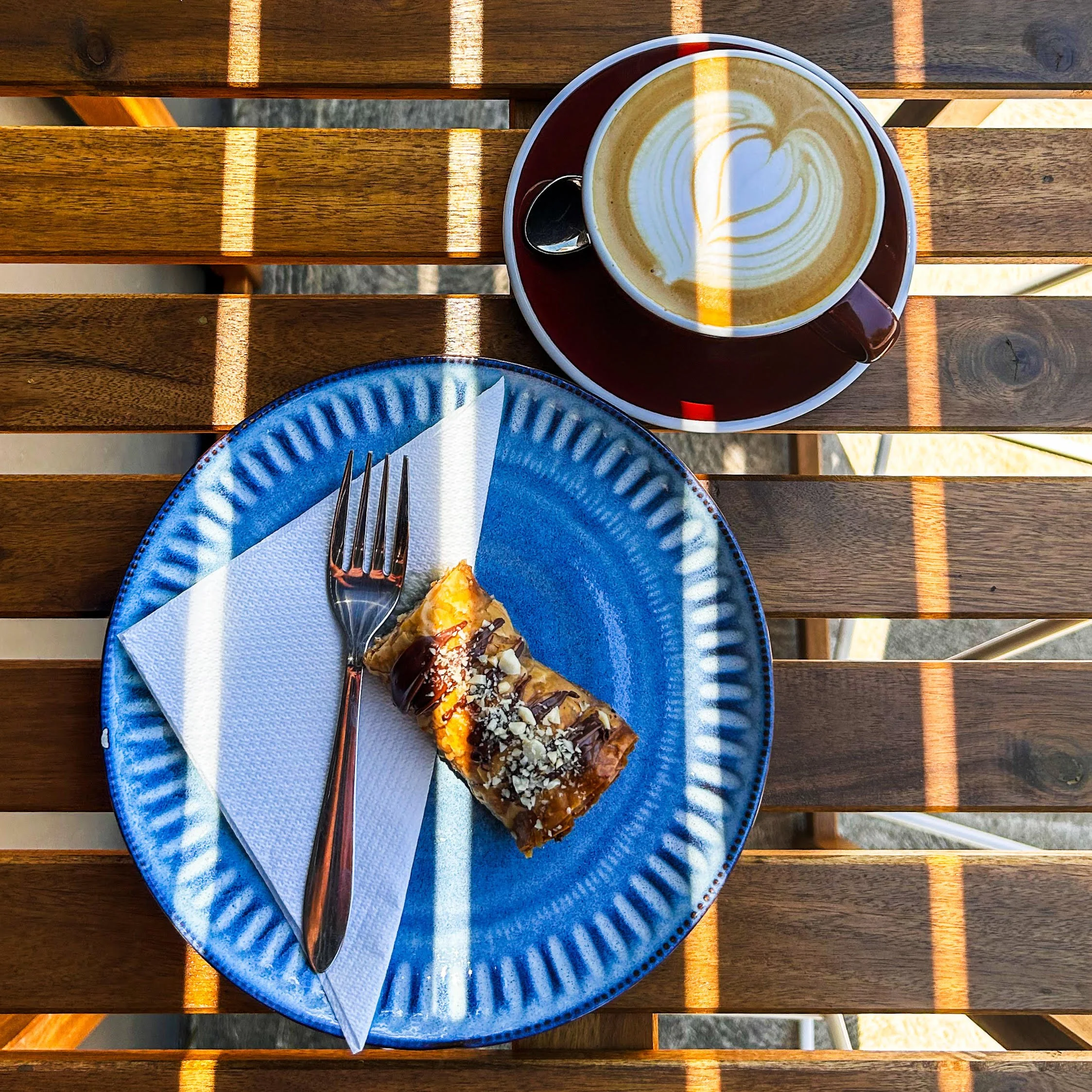 A cup of coffee with latte art on a wooden table. A dessert and a fork are on a blue plate with a napkin. Sunlight casts shadows on the table.