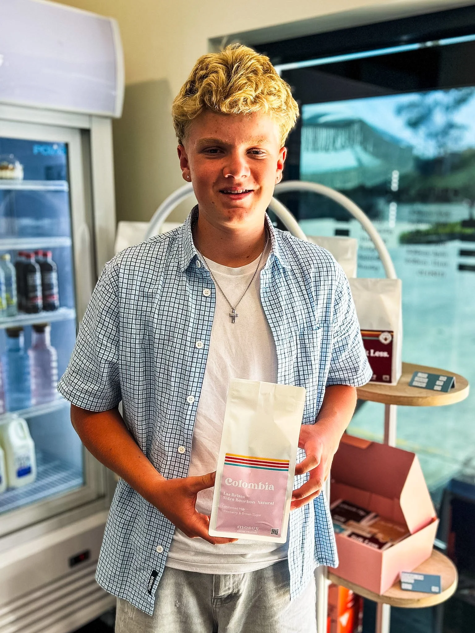 Young person with blond hair, wearing a checkered shirt, holding a pink promotional flyer for Colombia coffee, standing in front of a refrigerator and a drink display at a store or cafe.