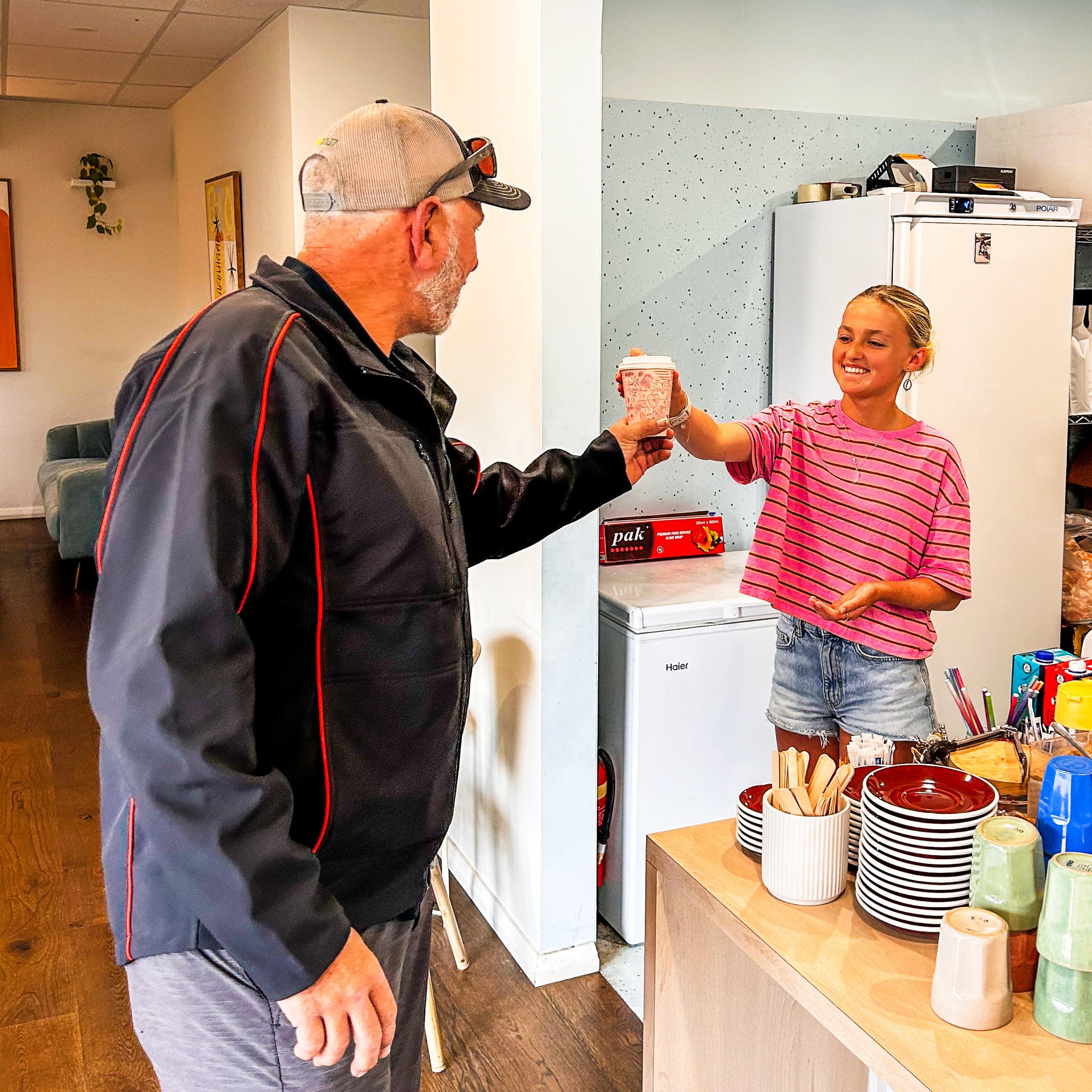 A young woman with blonde hair smiling and wearing a pink striped shirt is handing a hot drink in a decorated cup to an older man wearing a gray cap, black jacket, and glasses, in a café or restaurant setting with shelves of plates and cups.