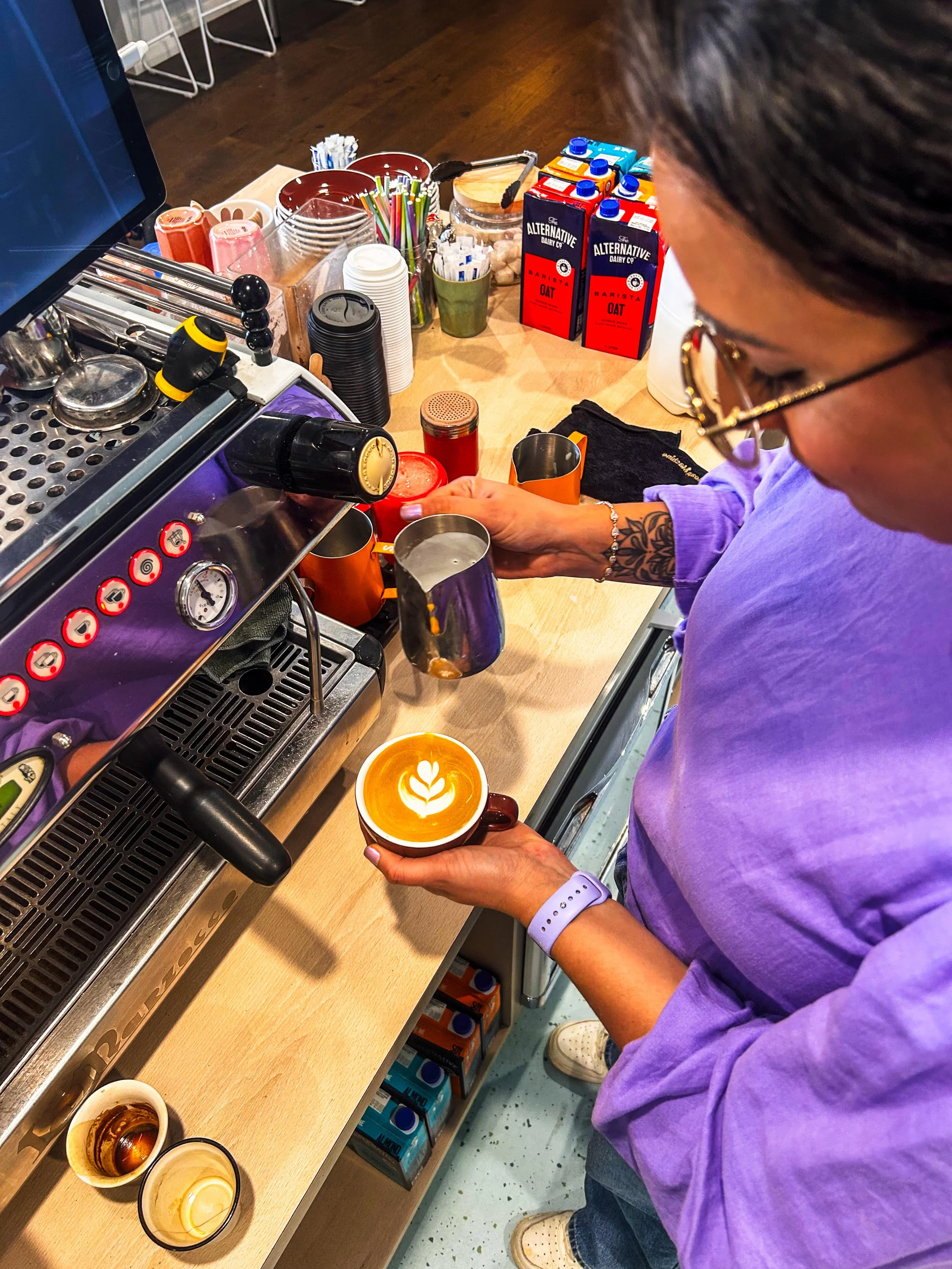 A barista preparing coffee behind a counter with various cups, cartons of oat milk, and utensils. A customer holds a cup of coffee with latte art.