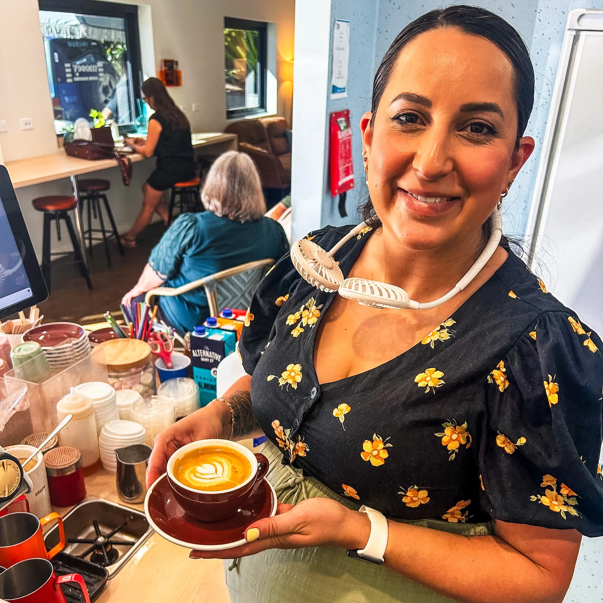 Woman in a black floral shirt holds a cup of coffee with latte art, standing in a cafe kitchen with various mugs, jars, and a stove in the background.