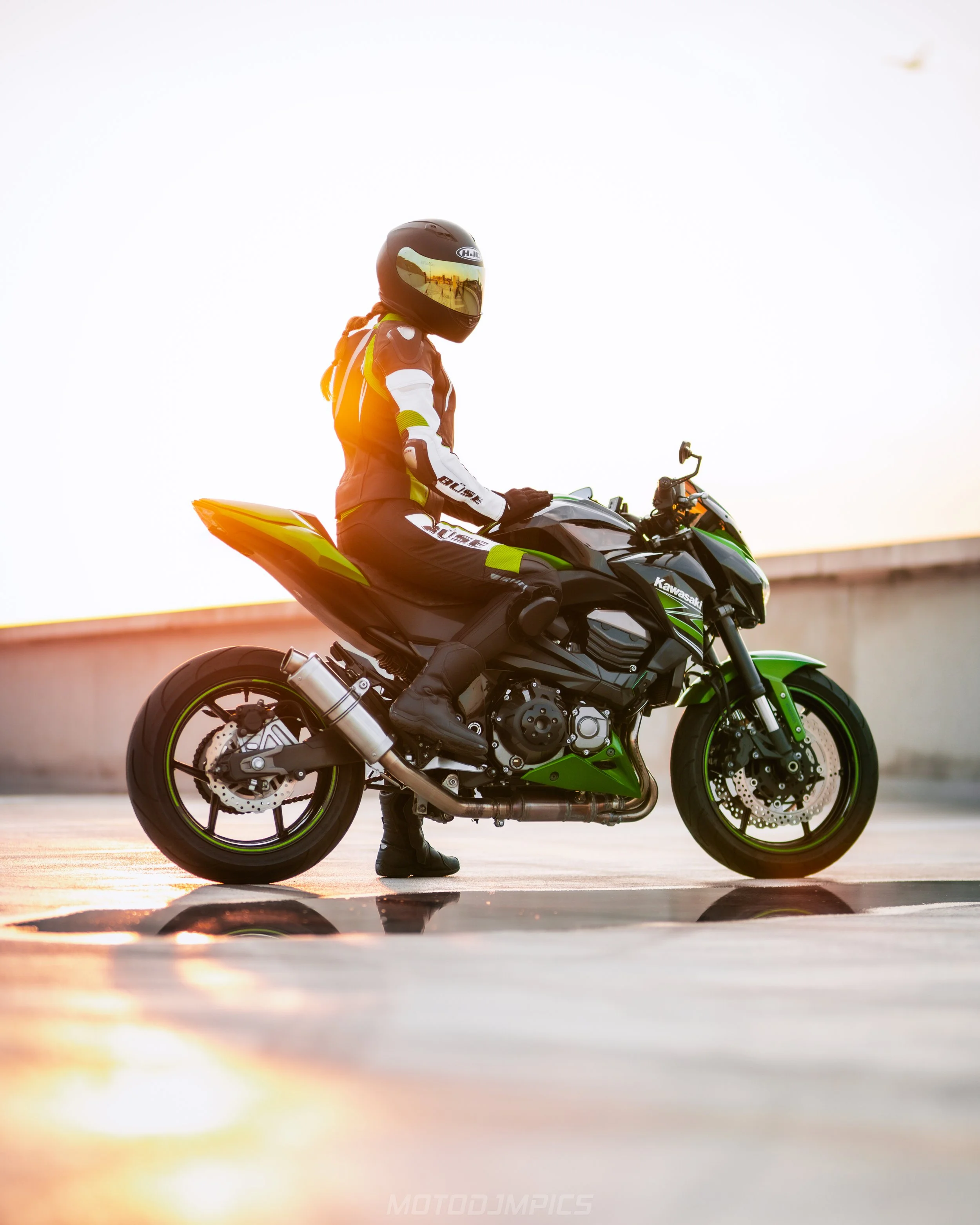 Persoon in motorpak en helm zittend op een groene en zwarte Kawasaki-motorfiets op een open weg met een lage zonsondergang op de achtergrond.
