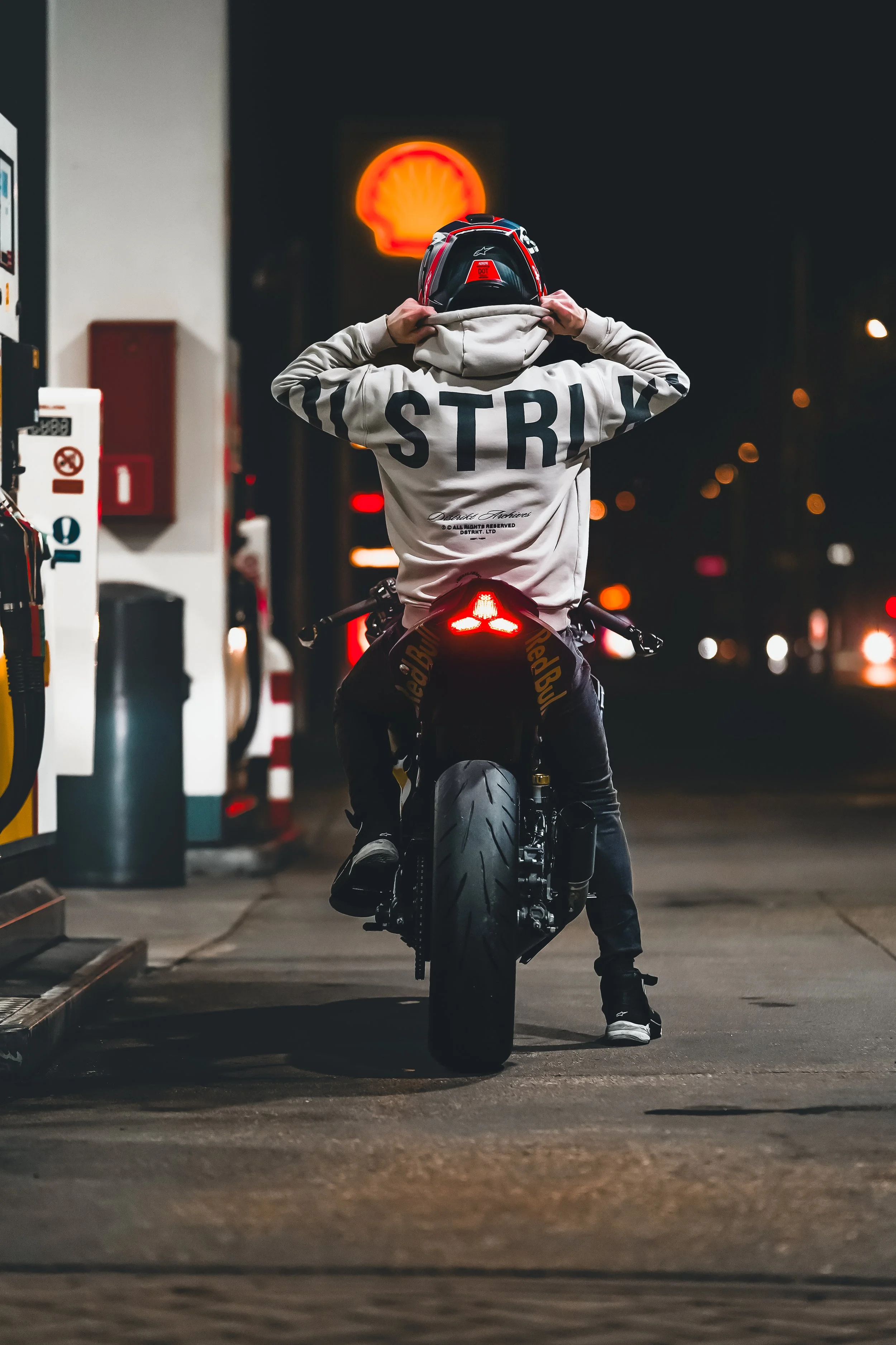 Persoon zit op een zwarte motorfiets bij een tankstation 's nachts, met een helm en een hoodie met 'STROLL' gedrukt op de achterkant, en het Shell-logo op de achtergrond.