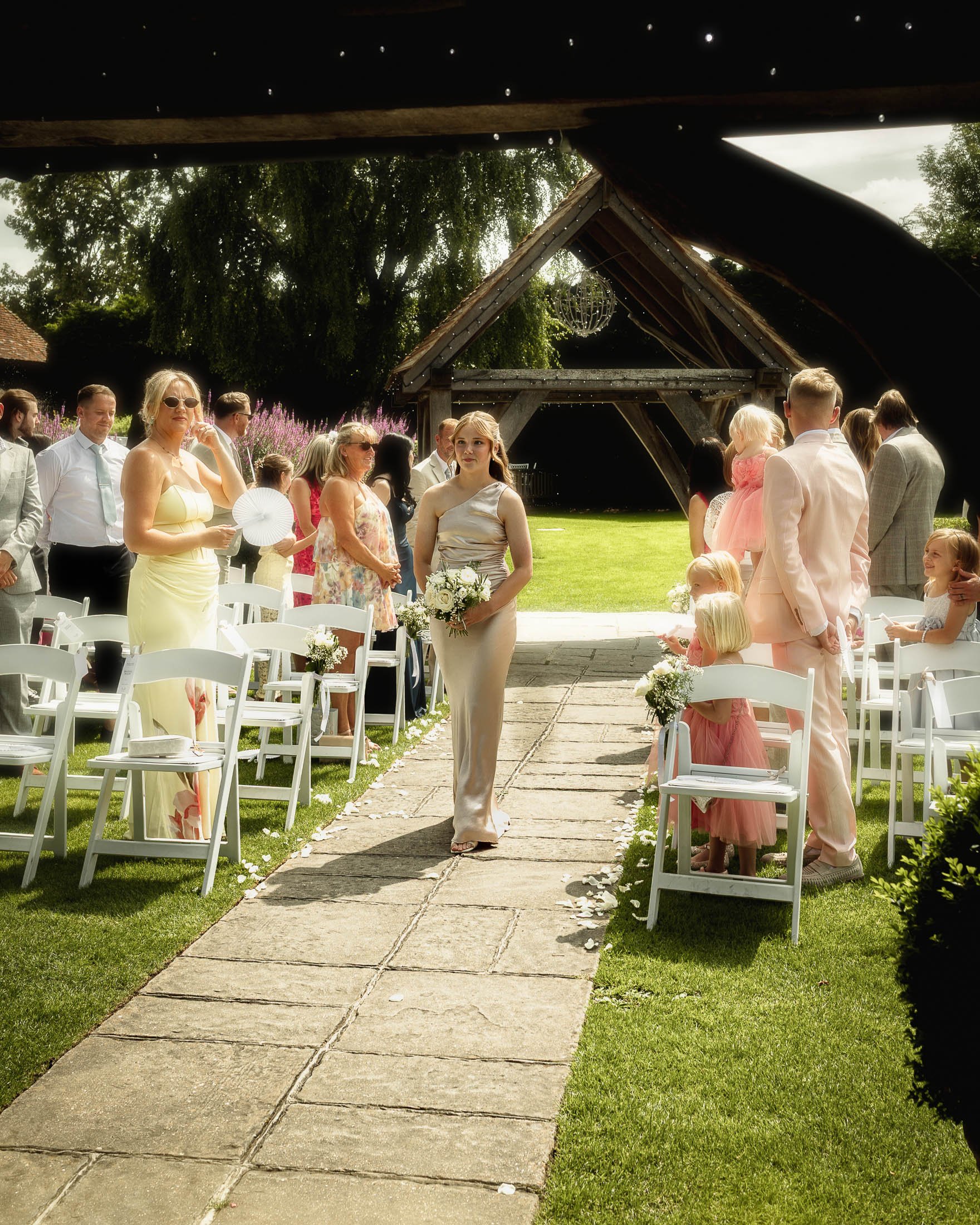 A bridesmaid wearing a champagne dress and holding a bouquet of white flowers walks down the aisle during an summer outdoor wedding ceremony at Winters Barns in Kent.