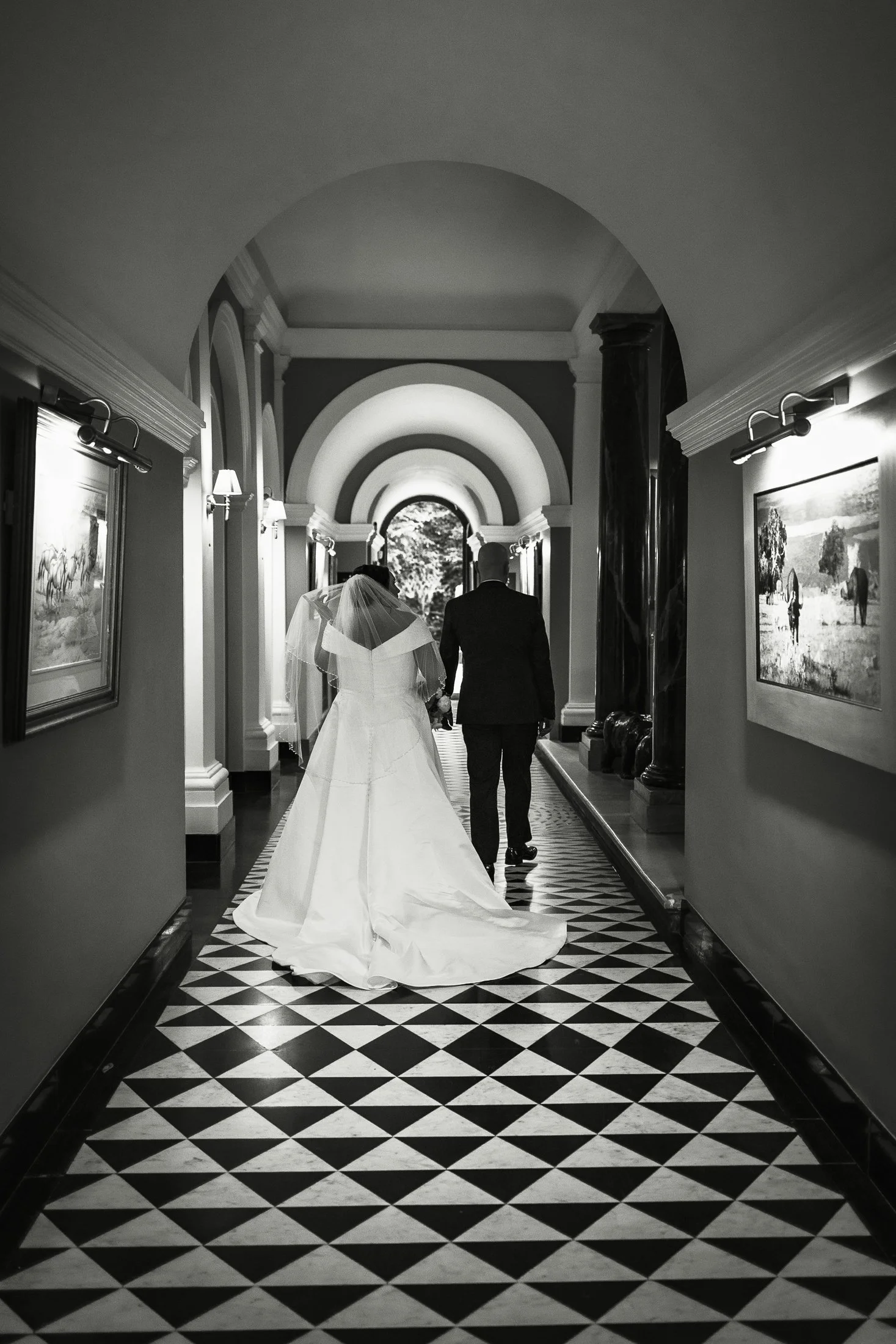 Black and white editorial image of a bride and groom walking through the internal main corridor of the Mansion at Port Lympne Hotel & Reserve.