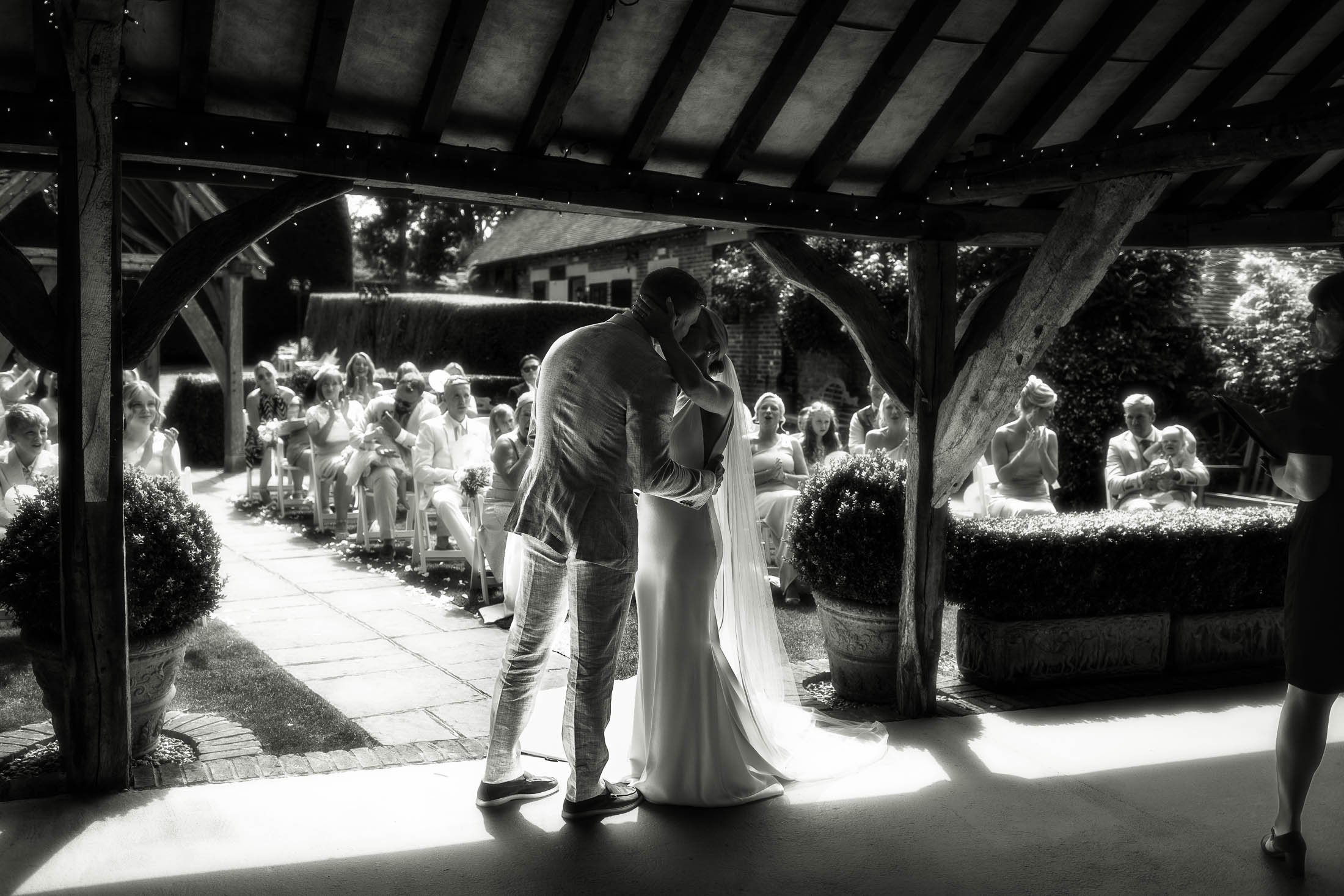 A couple share their first kiss during their outdoor wedding ceremony at Winters Barns in Canterbury, Kent, captured in a black and white timeless wedding photo aesthetic.
