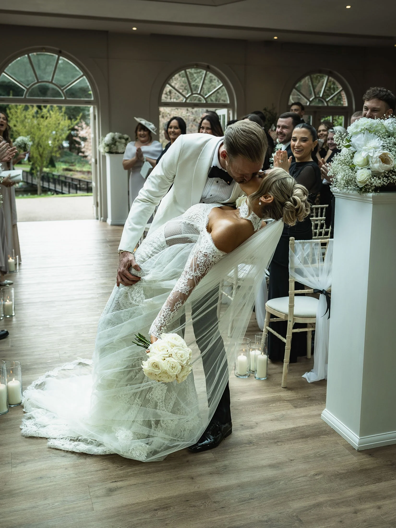 White tuxedo groom dips and kisses his bride as they exit their indoor wedding ceremony at the Orangery Maidstone in Kent.