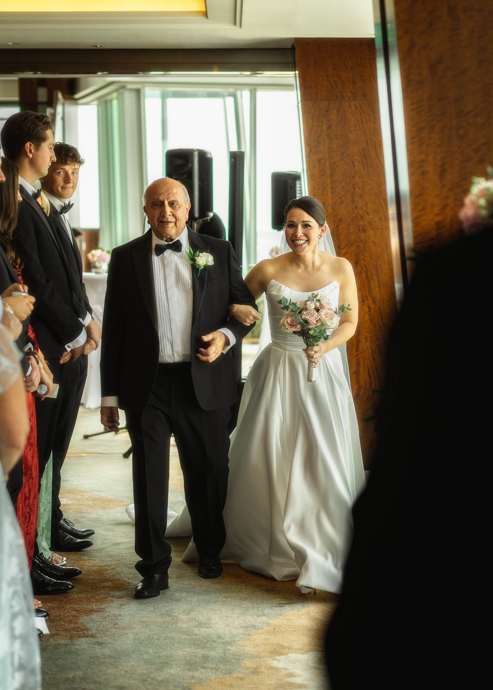 A bride walks arm-in-arm with her Father down the aisle of her indoor London wedding, a moment that is captured by a cohesive wedding photography and videography team.