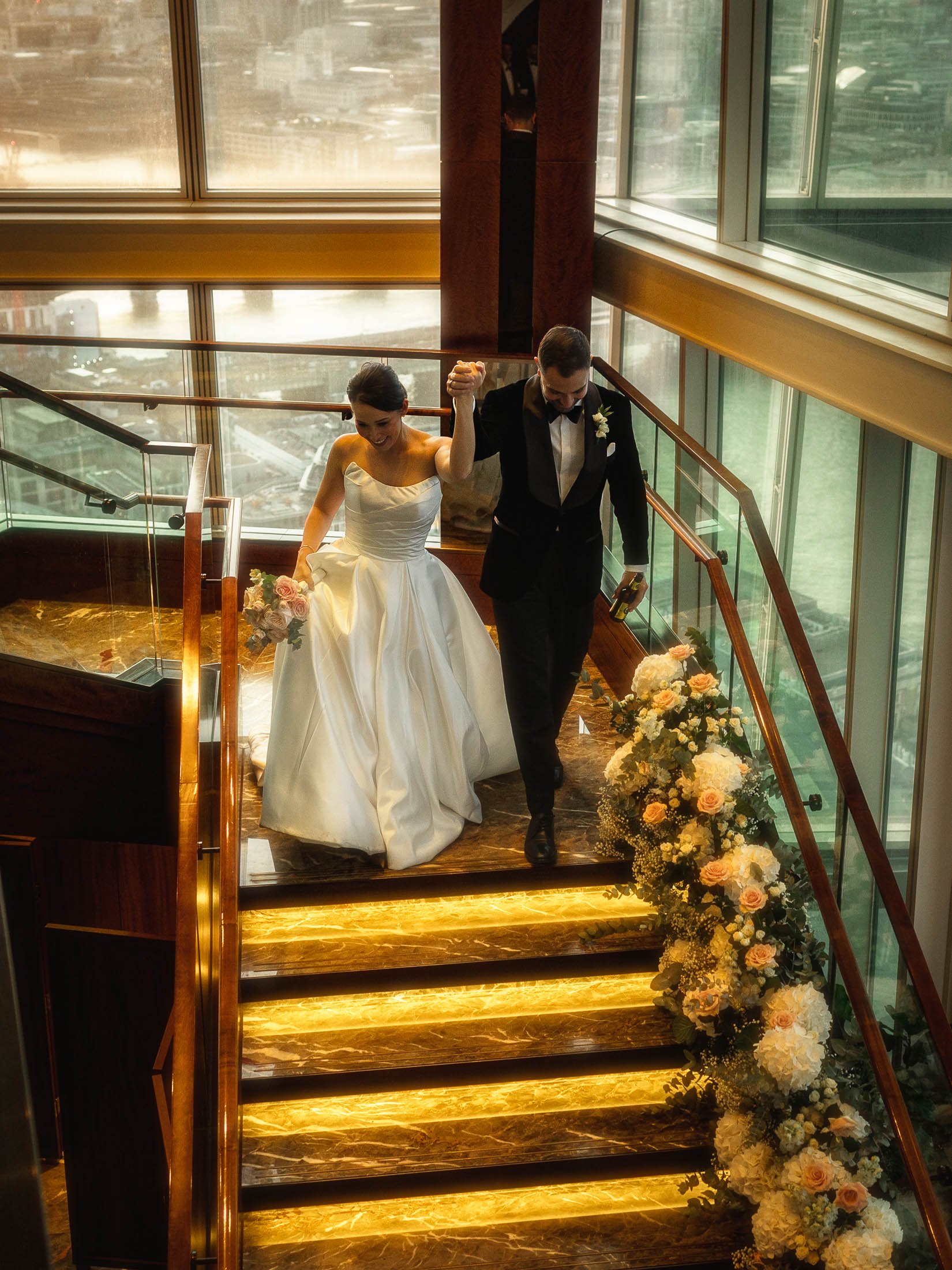 A bride and groom raise their hands in celebration as they join their black-tie wedding reception at the Shard and their guests all cheer.