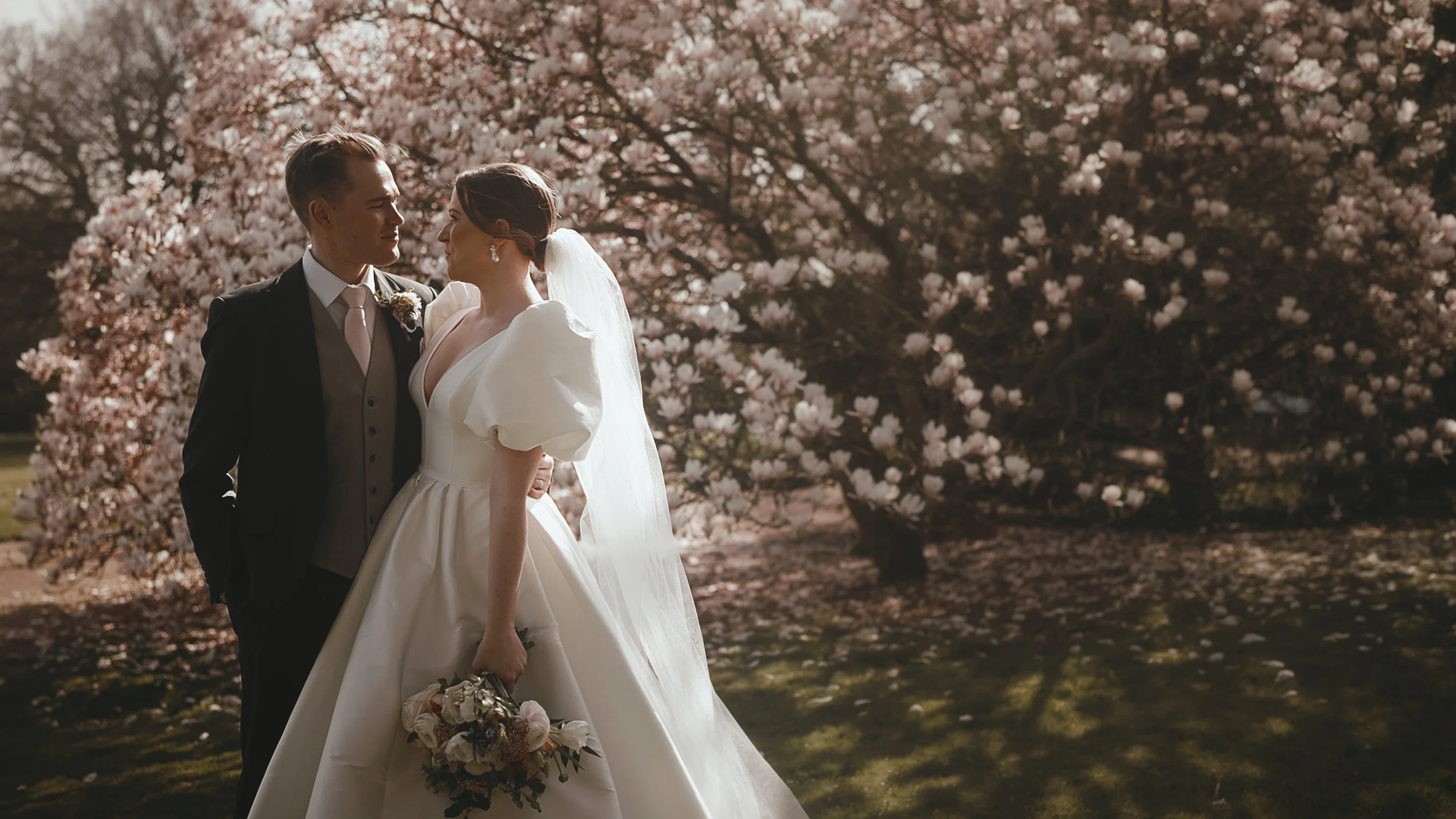 A bride and groom look at each other underneath a blossom tree at a Balcombe Place wedding in Sussex.