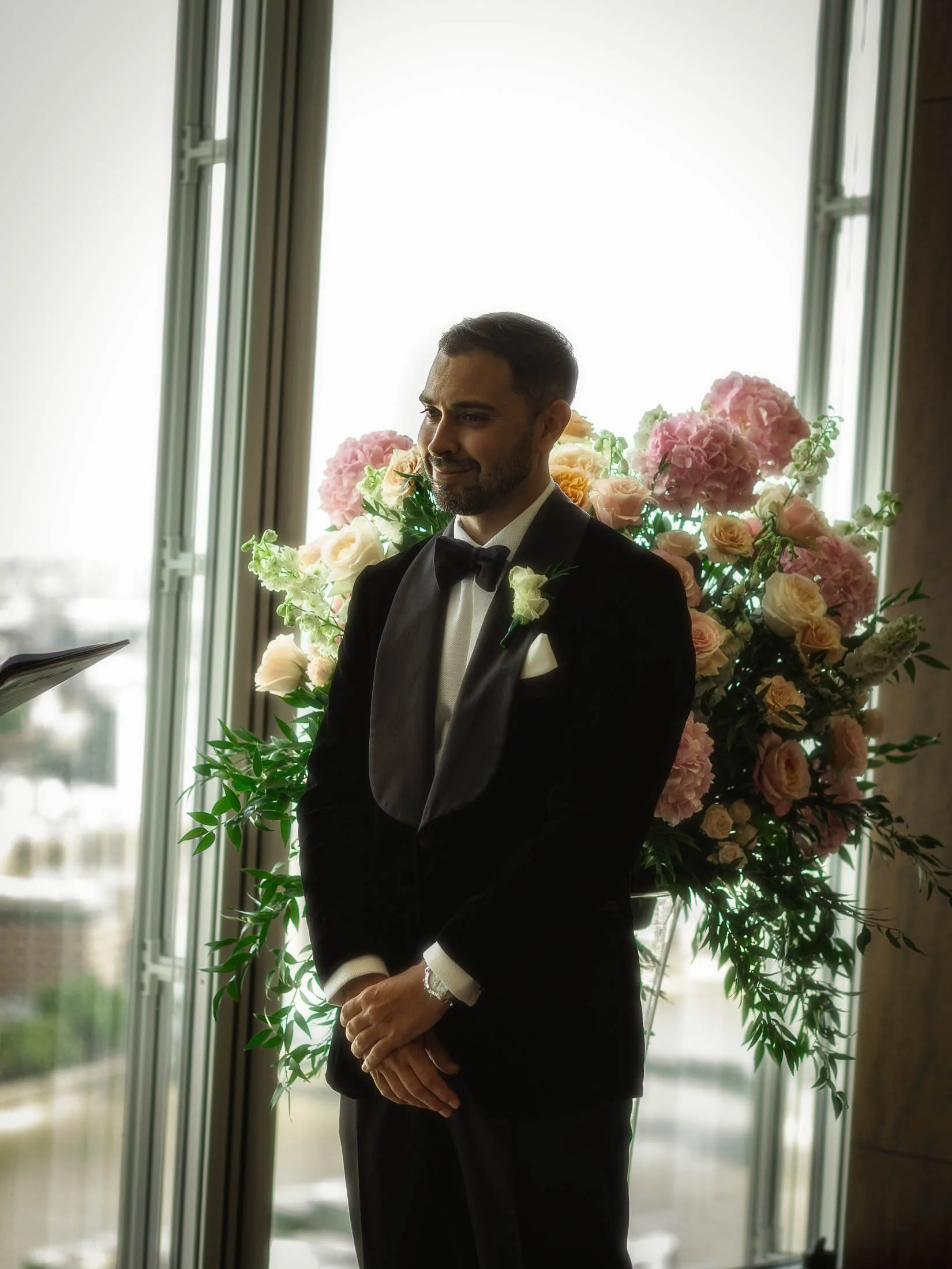 A groom in black tie tuxedo waits at the end of the aisle during an indoor wedding ceremony at the Shangri-La hotel in the Shard.
