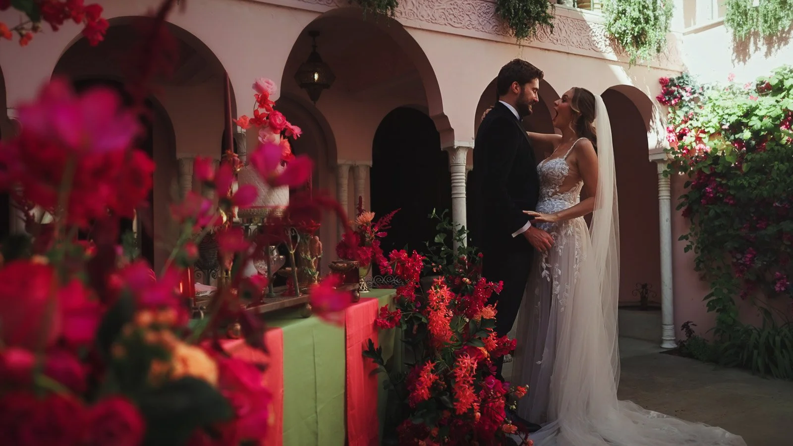 A couple stand amongst bright pink and red reception decor and florals on their colourful wedding day.