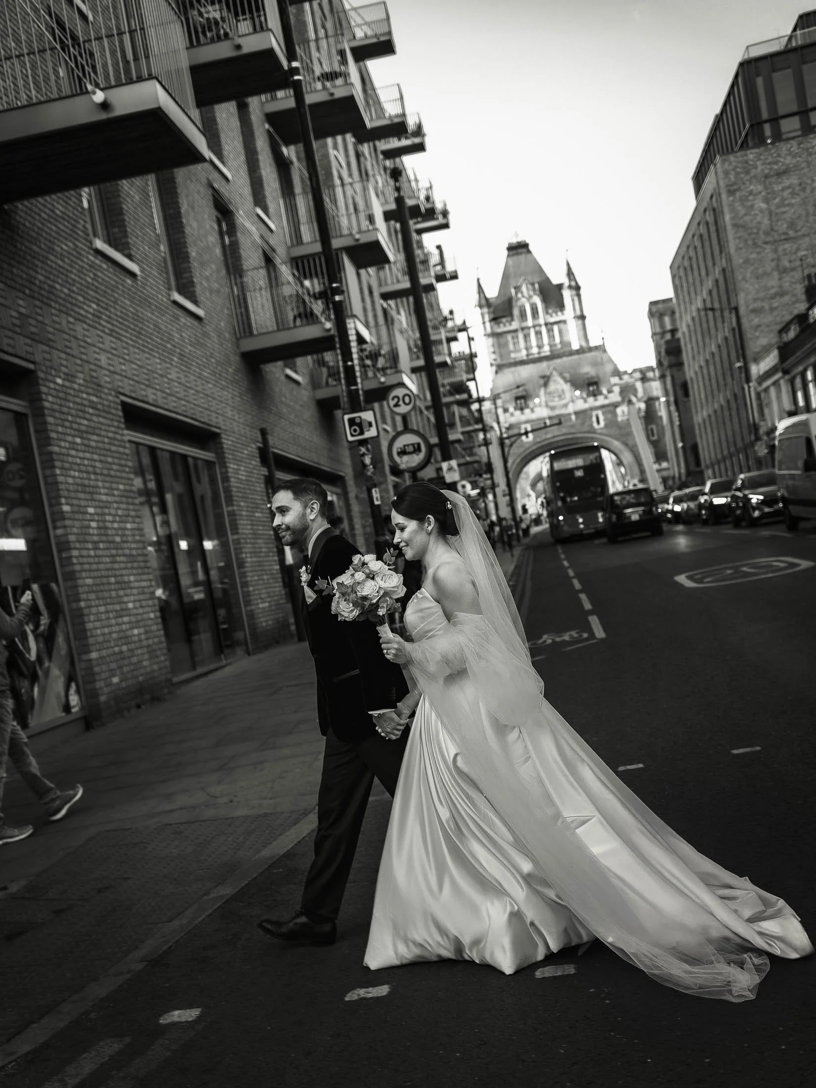 A natural yet editorial London wedding moment as a bride and groom cross a busy street by Tower Bridge during the black tie wedding portrait session on the streets of London.