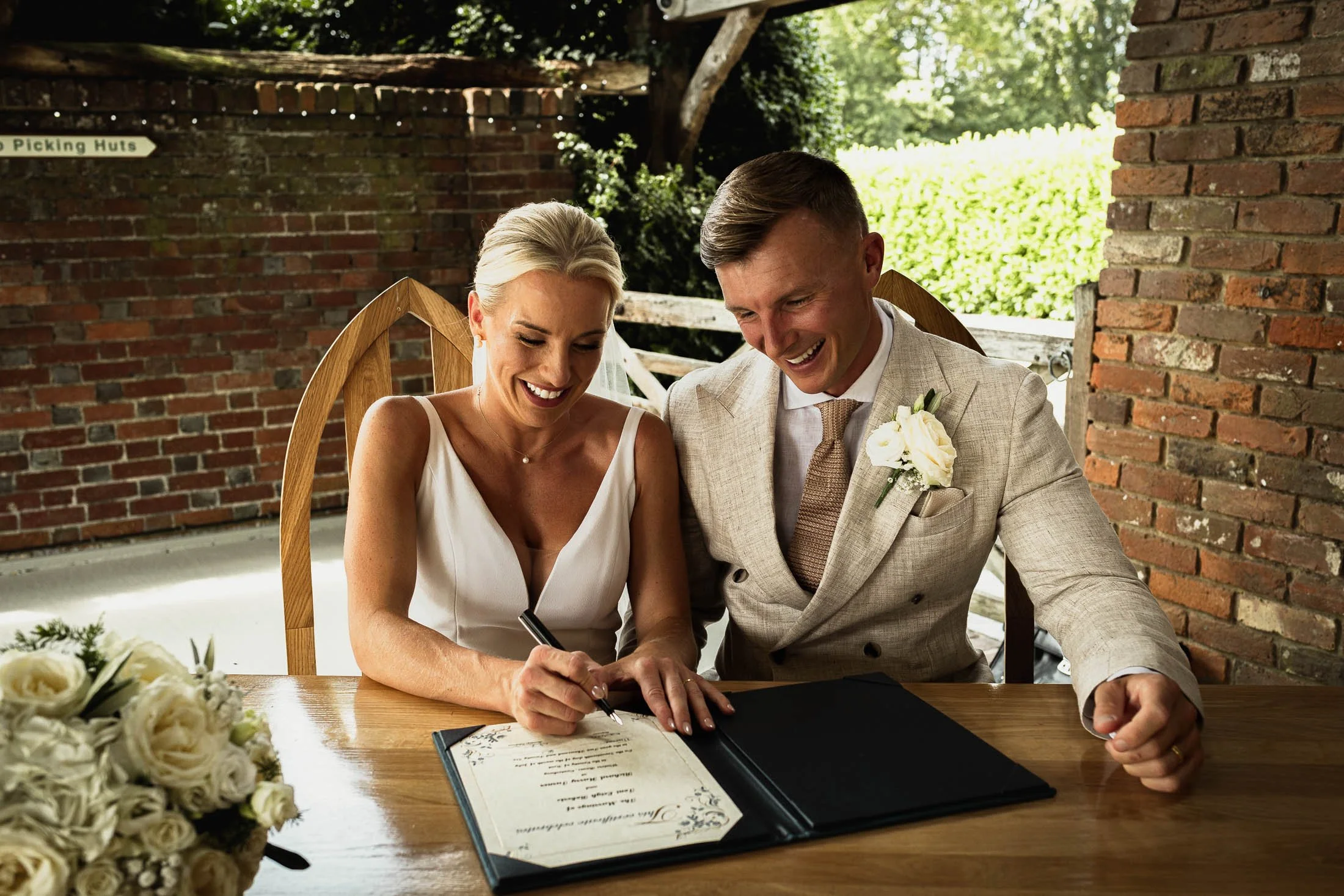 A bride and groom smiling together as they sign the register at the end of their outdoor wedding ceremony at Winters Barns in Kent.
