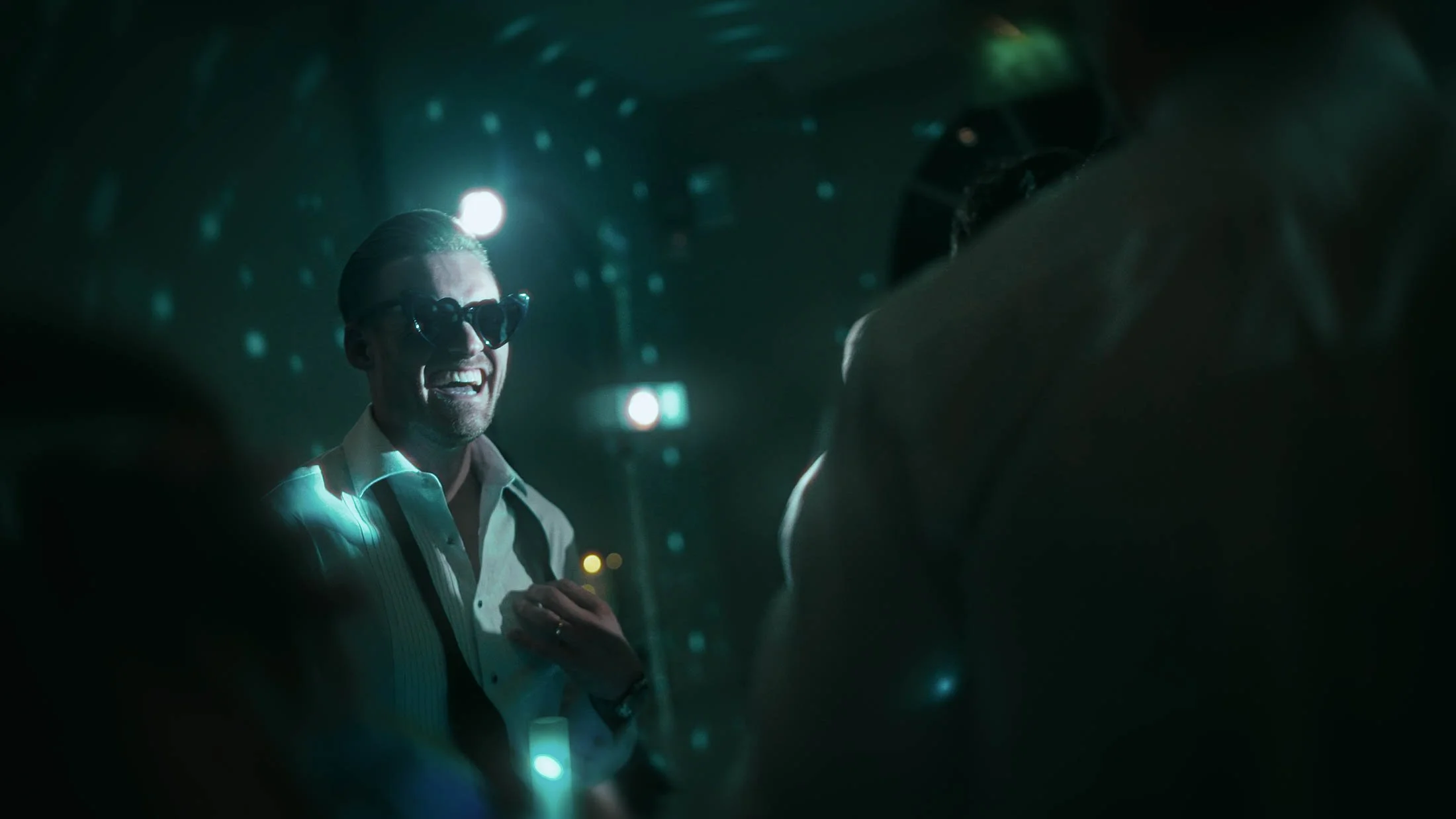 A groom dances with his guests during his wedding after party, with his bow tie undone and wearing heart-shaped sunglasses. Part of a high-energy sequence from a storytelling wedding film.