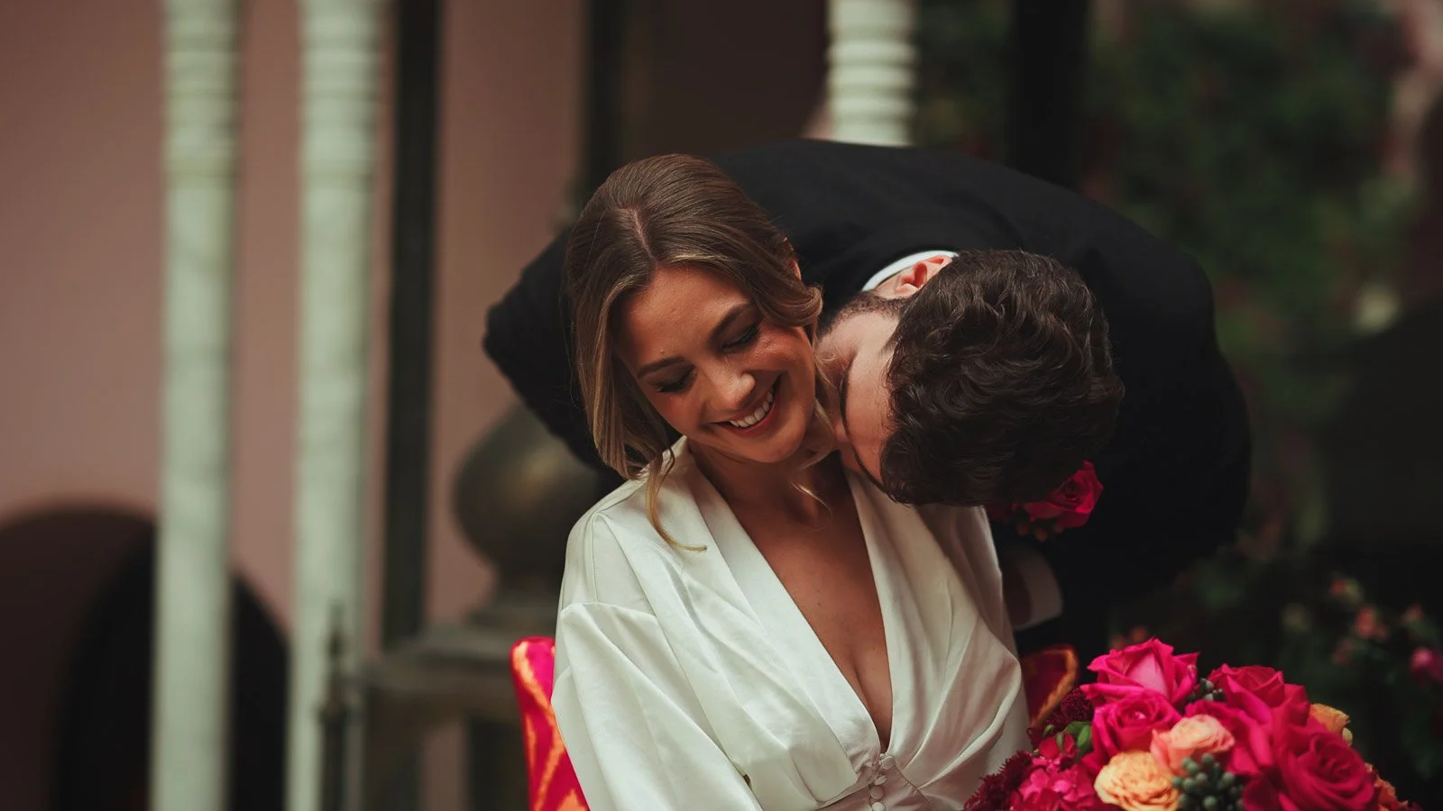 An intimate moment as a groom kisses his brides neck and she smiles, surrounded by bold pink and red florals.
