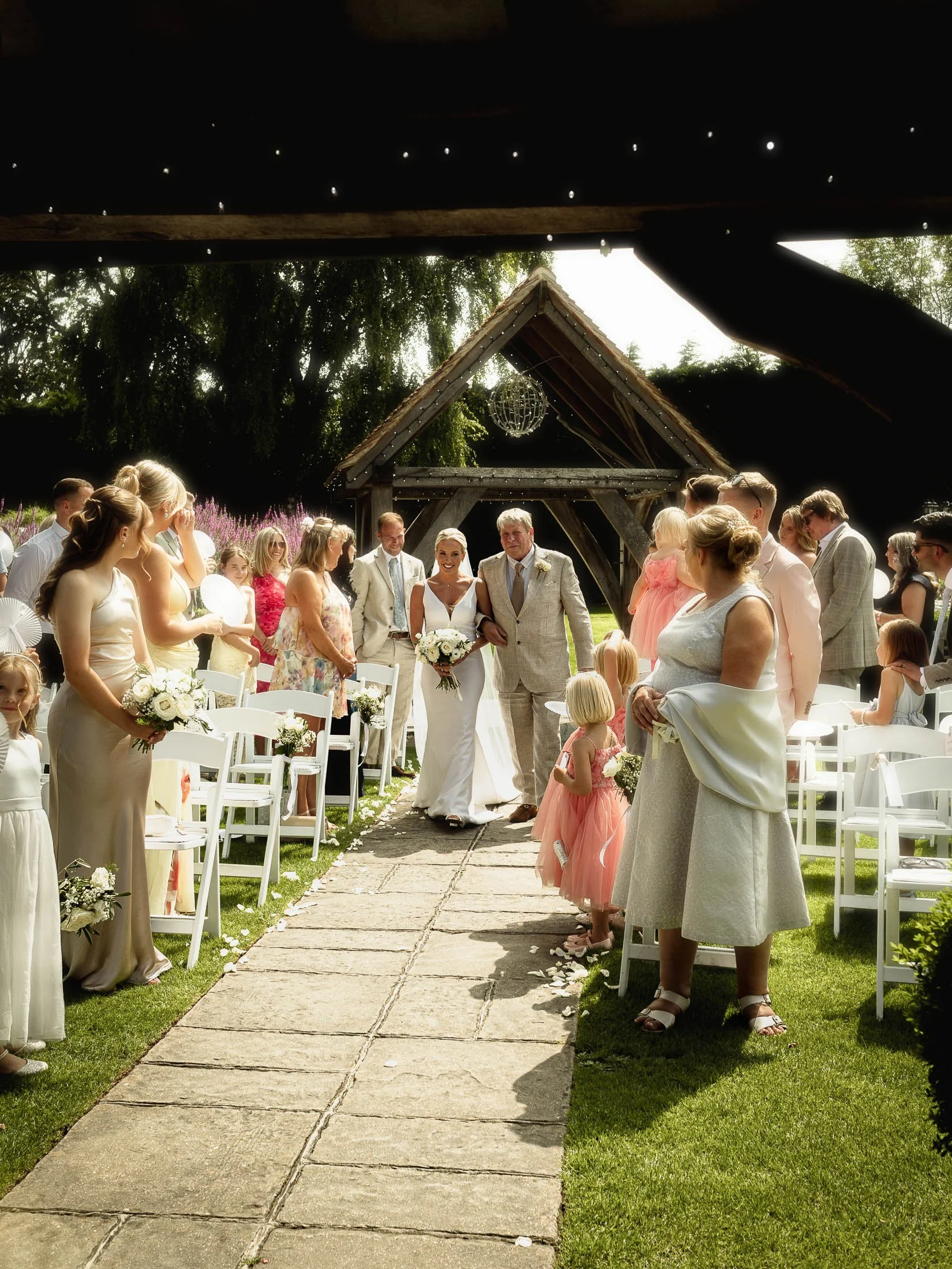 A bride walks arm-in-arm with her Father as she enters her summer outdoor wedding ceremony at Winters Barns, Kent. Captured by a wedding photography and videography team curated by Luke Batchelor Productions