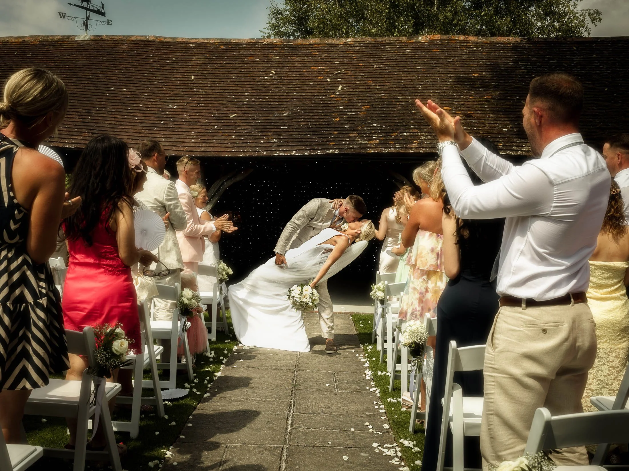 A groom dips and kisses his bride as they exit their colourful summer outdoor wedding ceremony at Winters Barns in Kent, as they are clapped and cheered by their wedding guests.