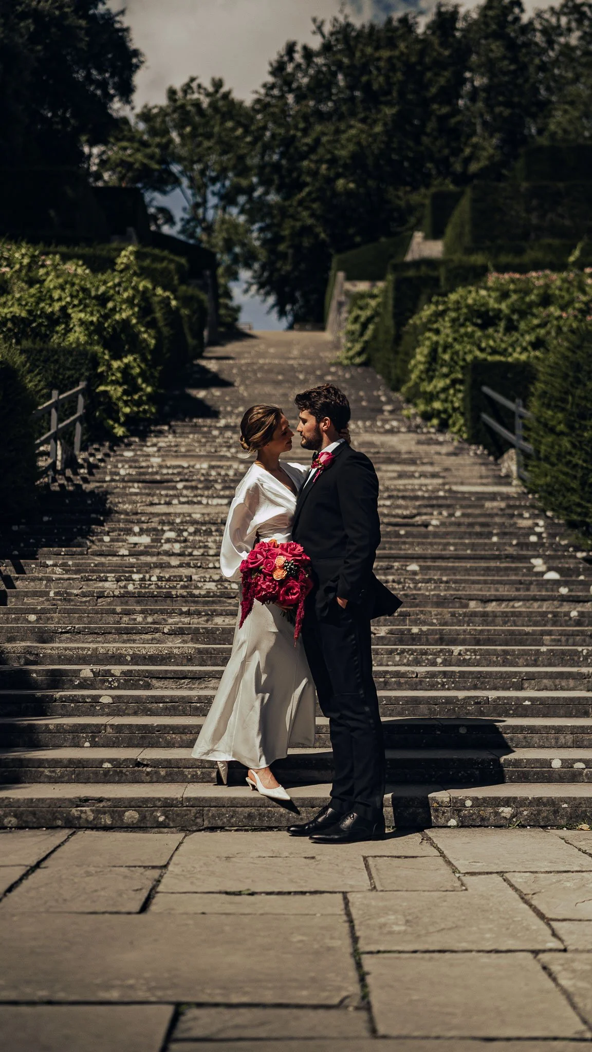 Couple portrait beneath trees, dappled light and fine art restraint at the bottom of a long stone staircase