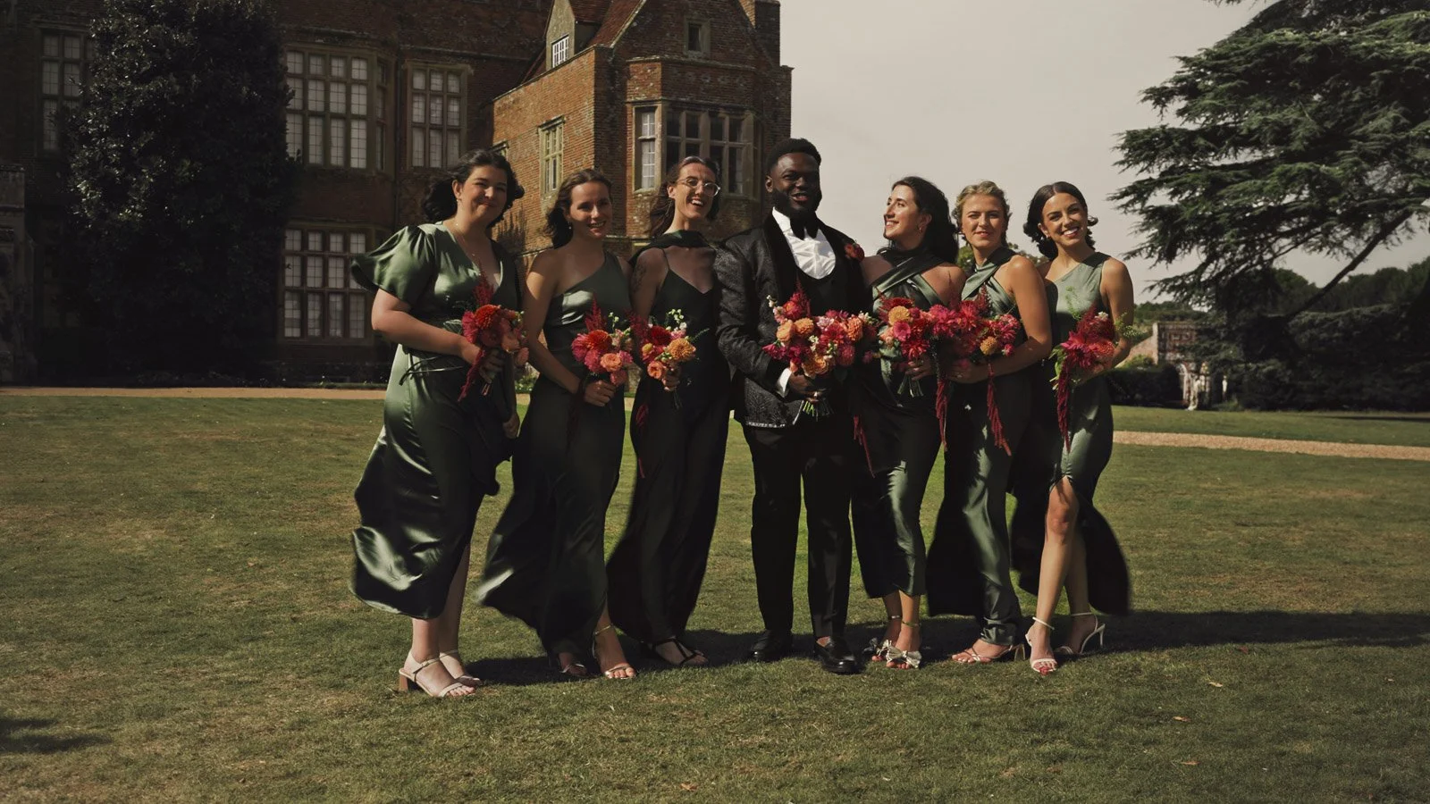 A groom in black tie stands with bridesmaids wearing green dresses, with all holding bright pink and orange bouquets during a colourful riverside wedding.