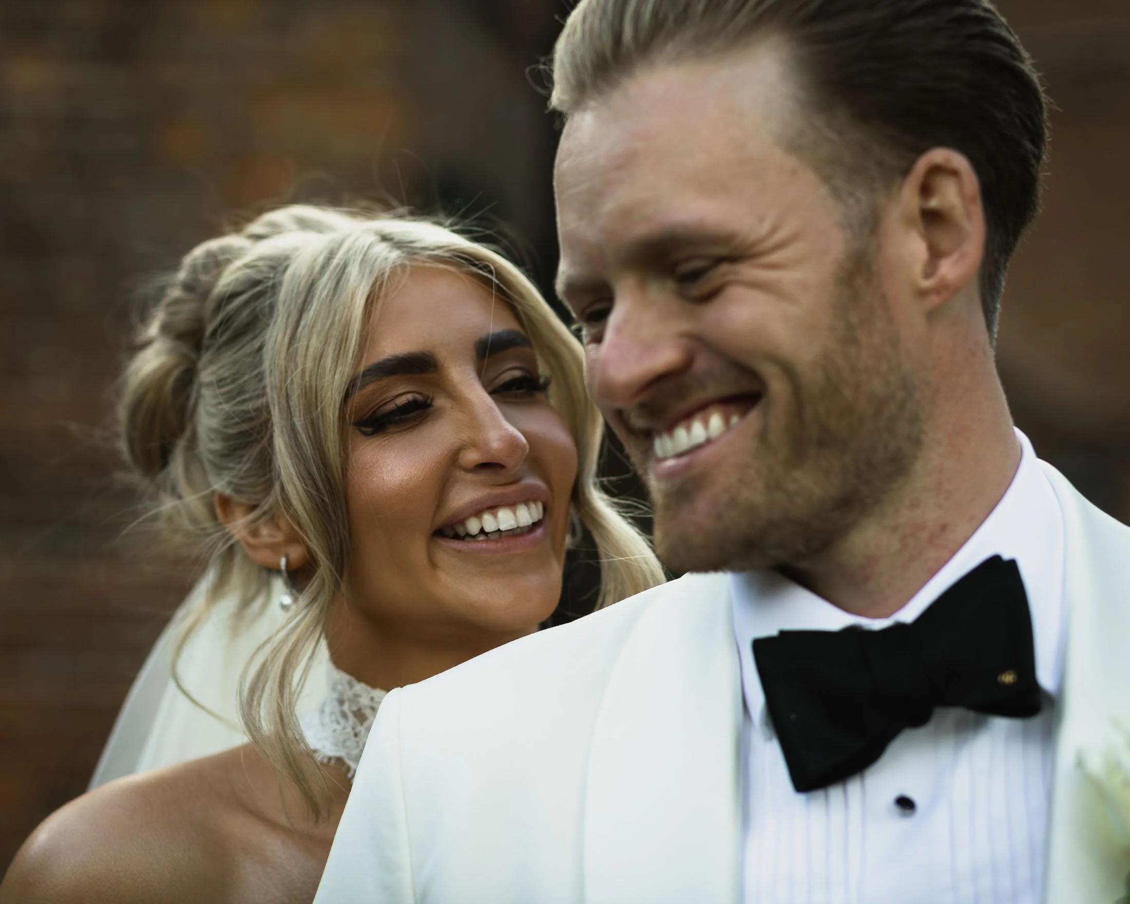A close up shot in an editorial composition of a bride smiling at her groom in a white tuxedo at the Orangery Maidstone. Part of a testimonial that reads "We just watched the little trailer and cried. Wow wow wow I want to do it all again"