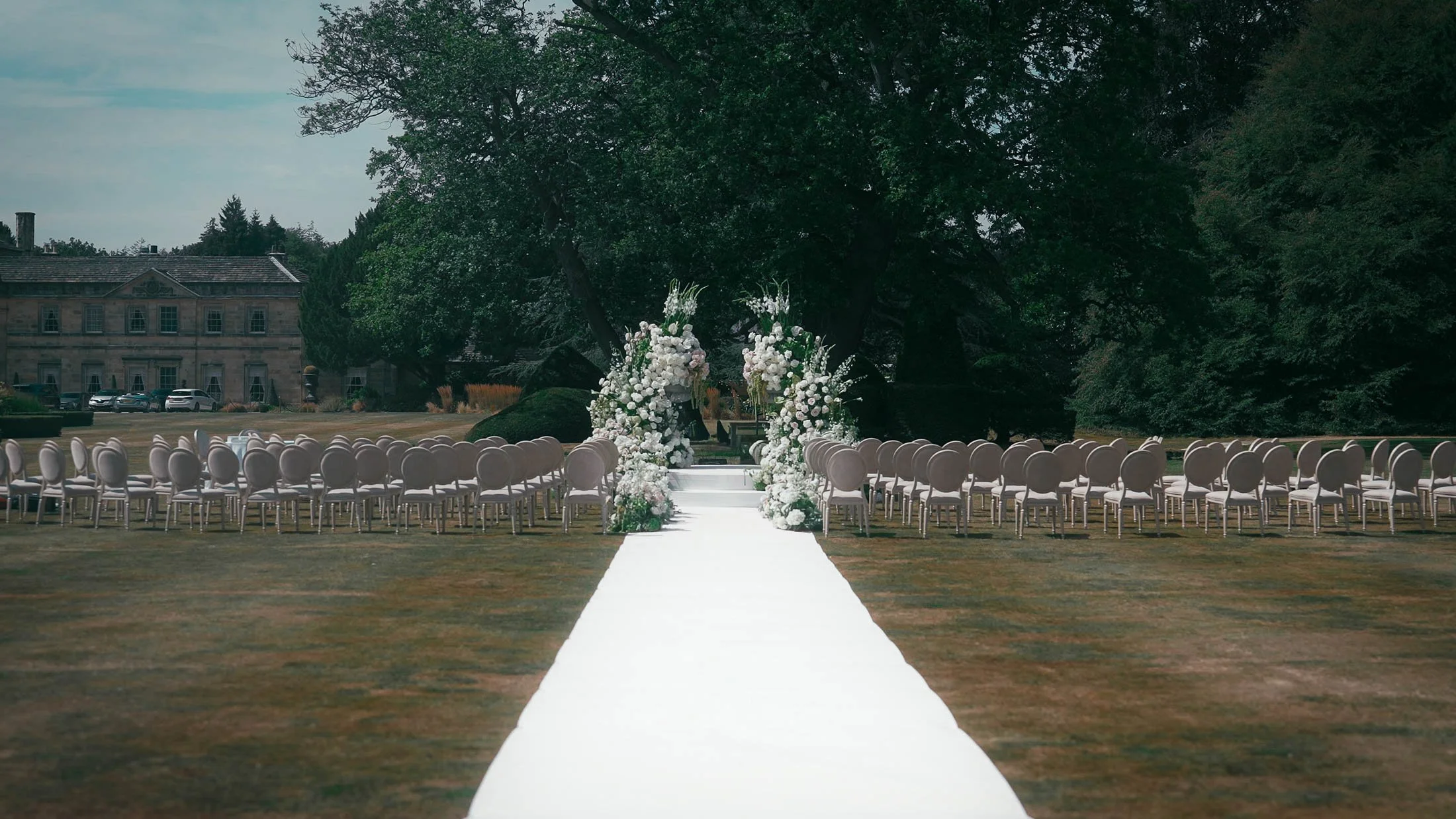 A Grantley Hall wedding videographer shot showing an outdoor wedding ceremony setup on the Front Lawn, with large white florals and a white ceremony aisle runner.