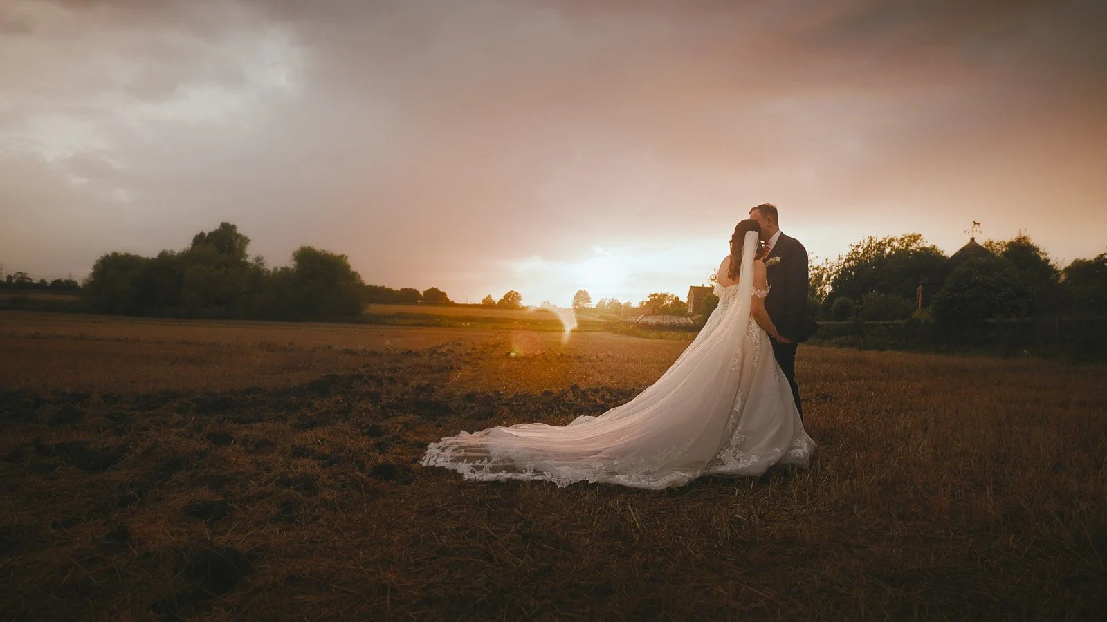 A bride and groom standing close together in a field at sunset, with the bride wearing a long white wedding gown and the groom in a dark suit, sharing a romantic moment.