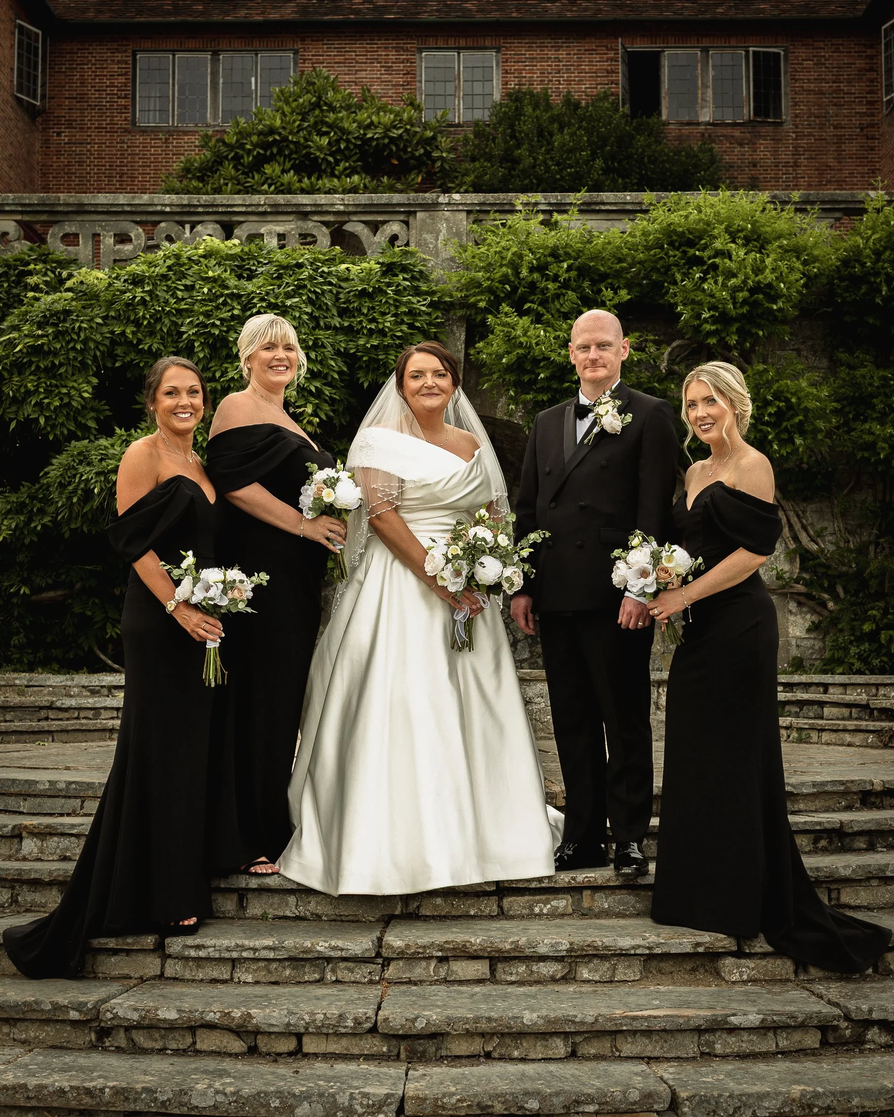 Bride Lucy stands with her bridesmaids in black dresses on the Mansion steps at port Lympne in Kent.
