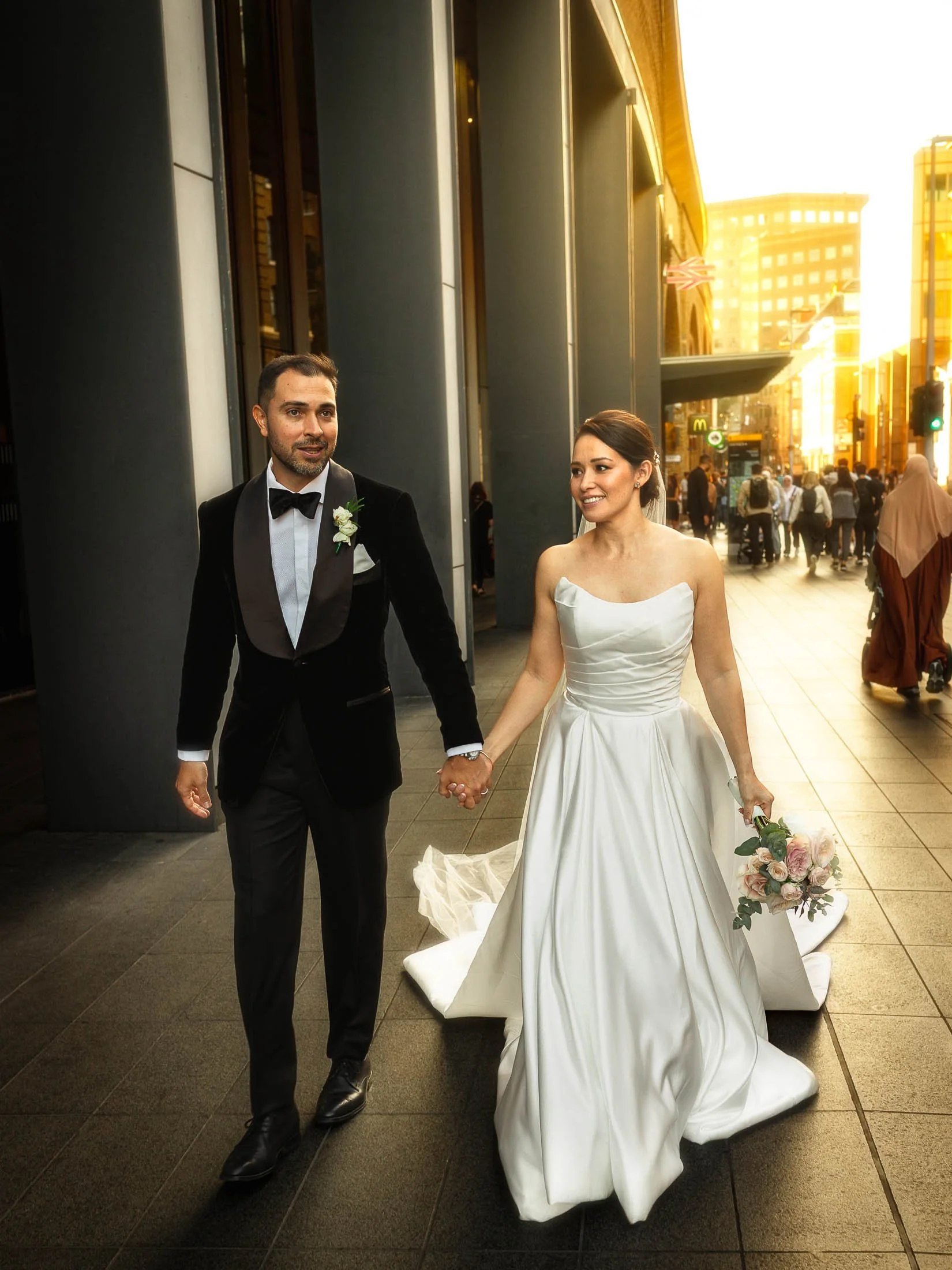 A groom in black tie tuxedo and a bride hold hands and smile as they walk through the streets of London during their modern black tie London wedding.