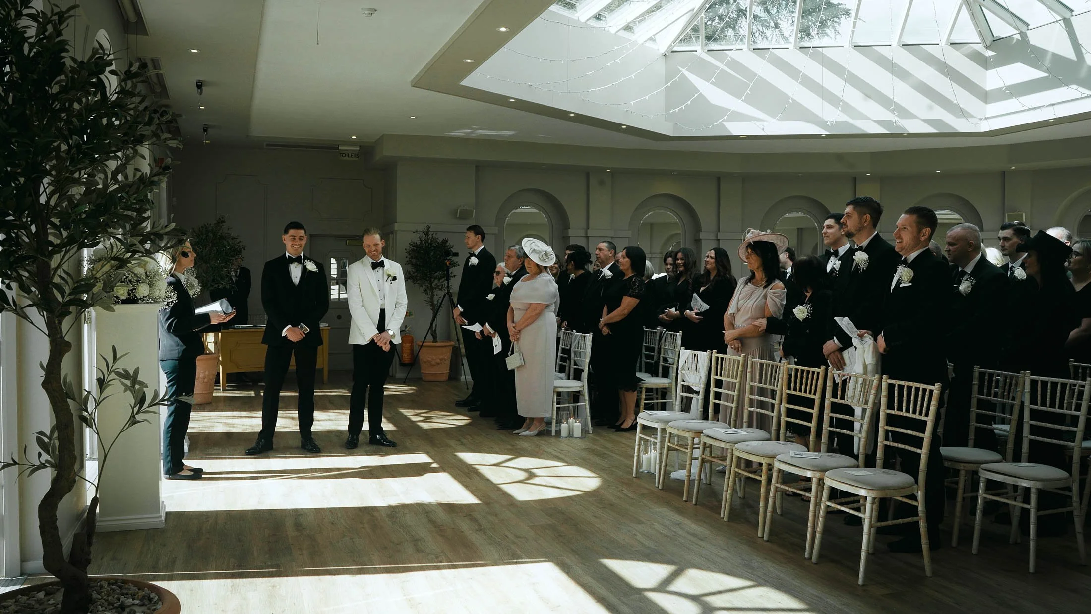A scene from a storytelling wedding film in the UK where a groom in black-tie waits at the front of the indoor ceremony for his bride to arrive.