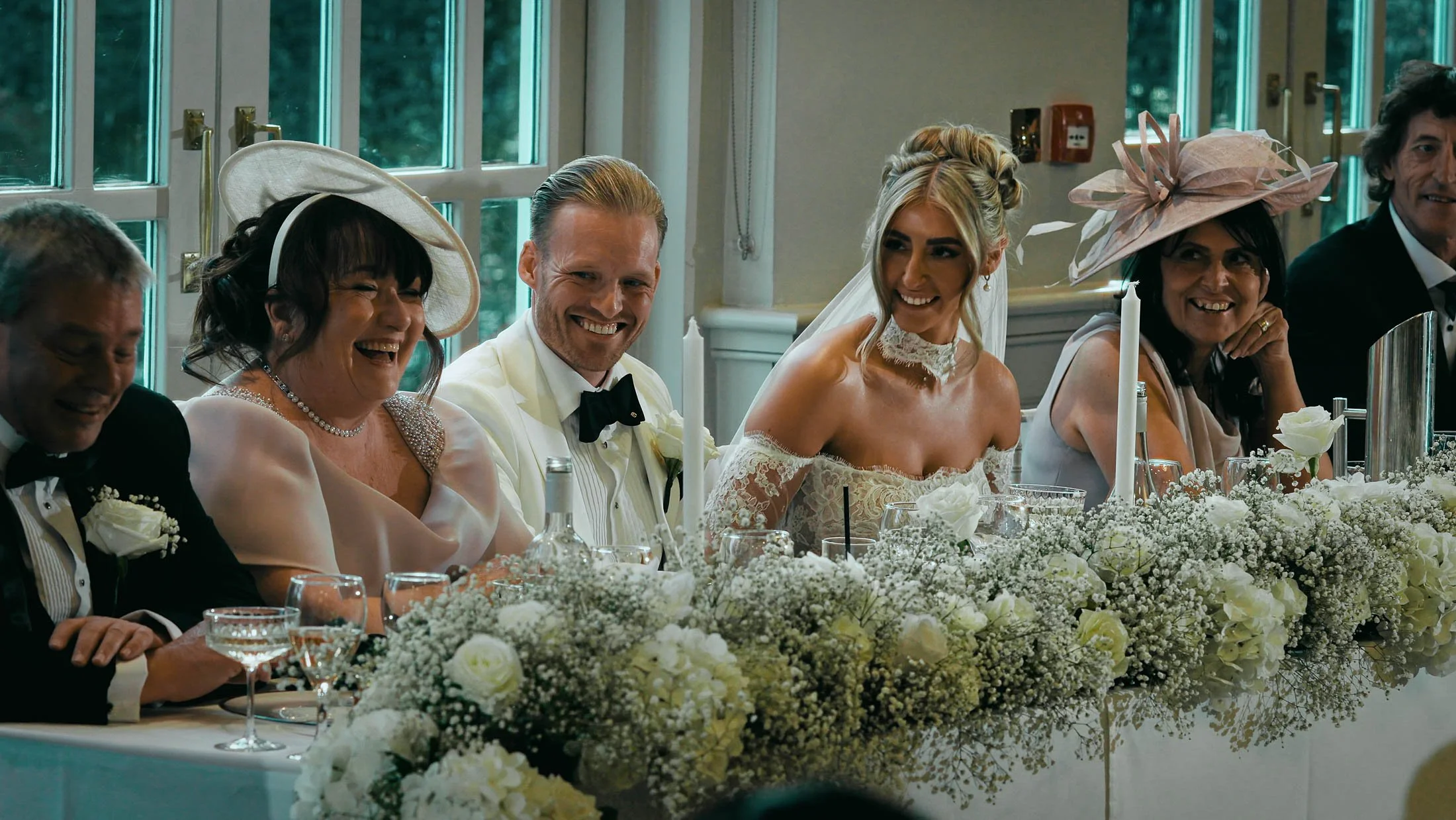 A bride and groom sit at the top table with their parents laughing during wedding speeches at The Orangery, Maidstone. Part of a storytelling cinematic wedding film captured in Kent, UK.