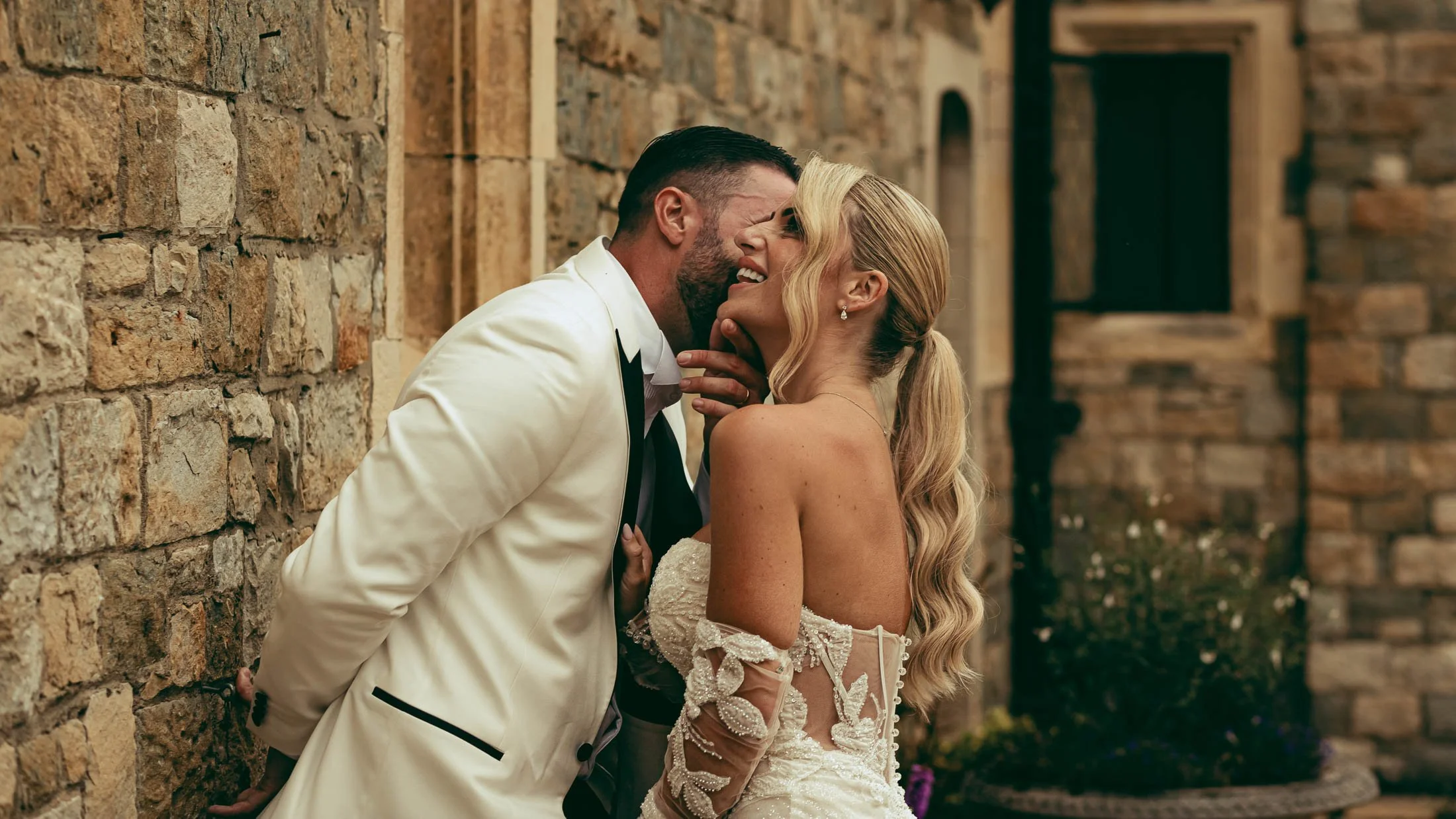 A fine art white tuxedo Eastwell Manor wedding in Kent, as a couple laugh together in a courtyard