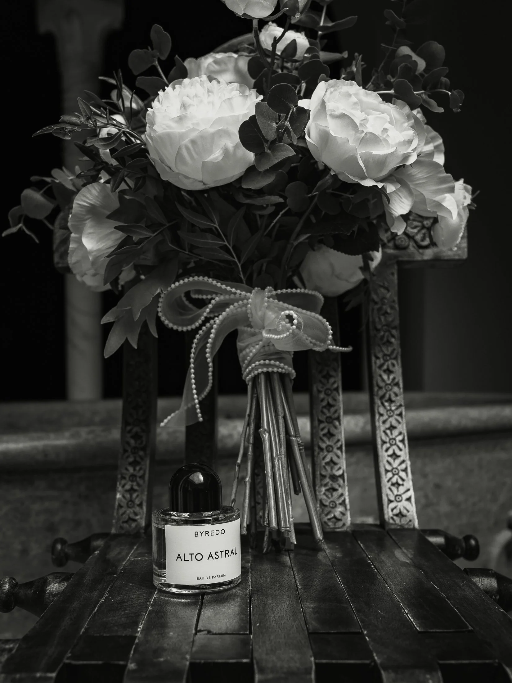 A black and white editorial wedding photograph showing a brides perfume bottle and bouquet on a wooden chair.
