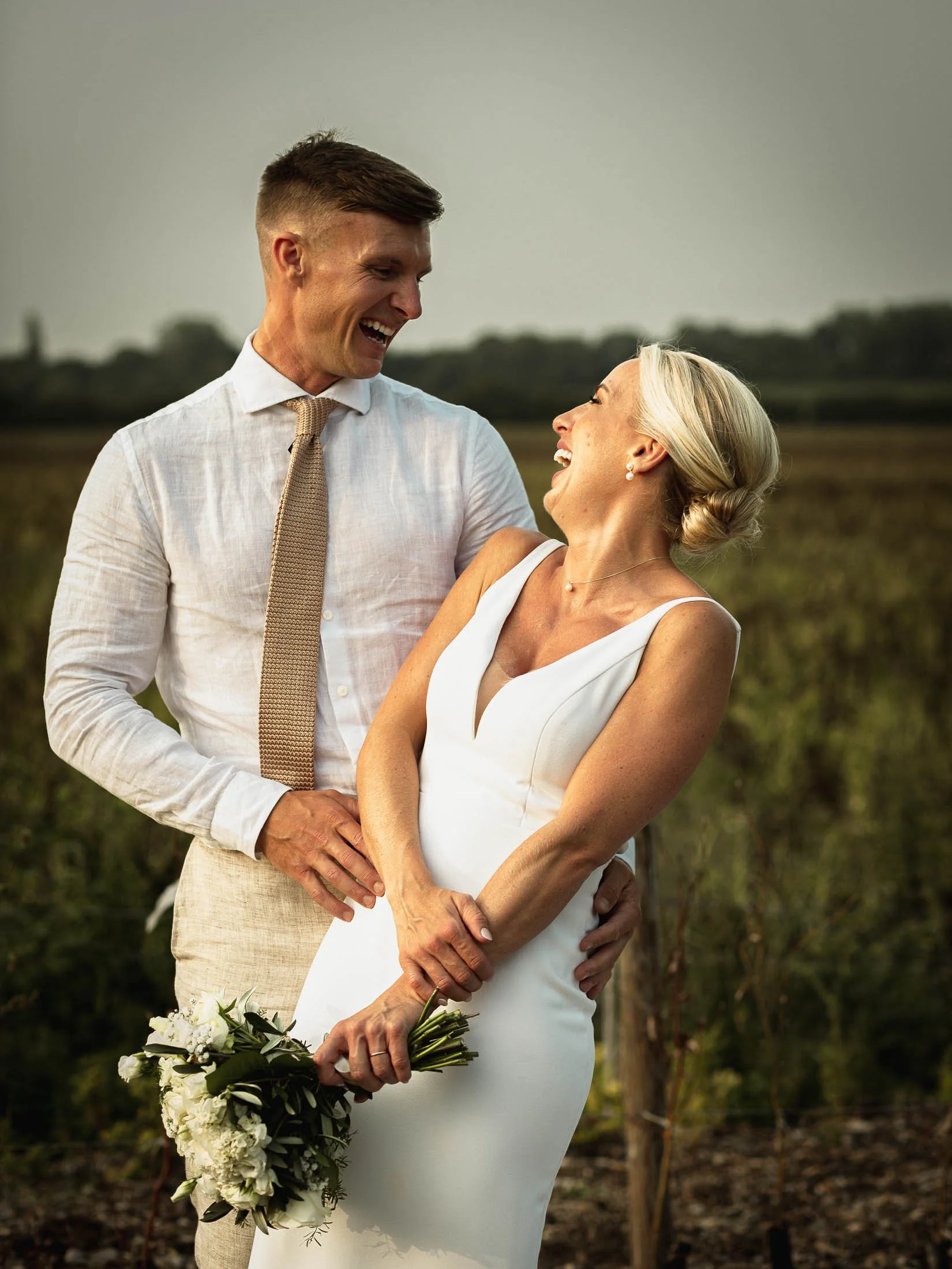 A bride and groom laughing heartily together during their golden hour portrait shoot at Winters Barns in Kent during an outdoor summer wedding day.
