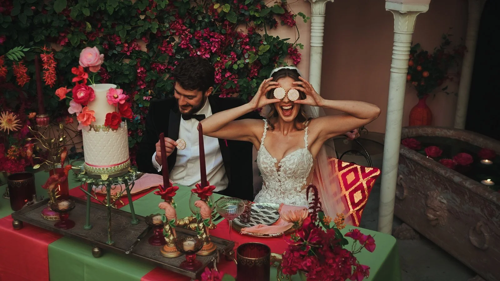 A candid editorial moment as a bride holds cookies over her eyes whilst sitting at a table decorated in bold pink wedding decor and accents.