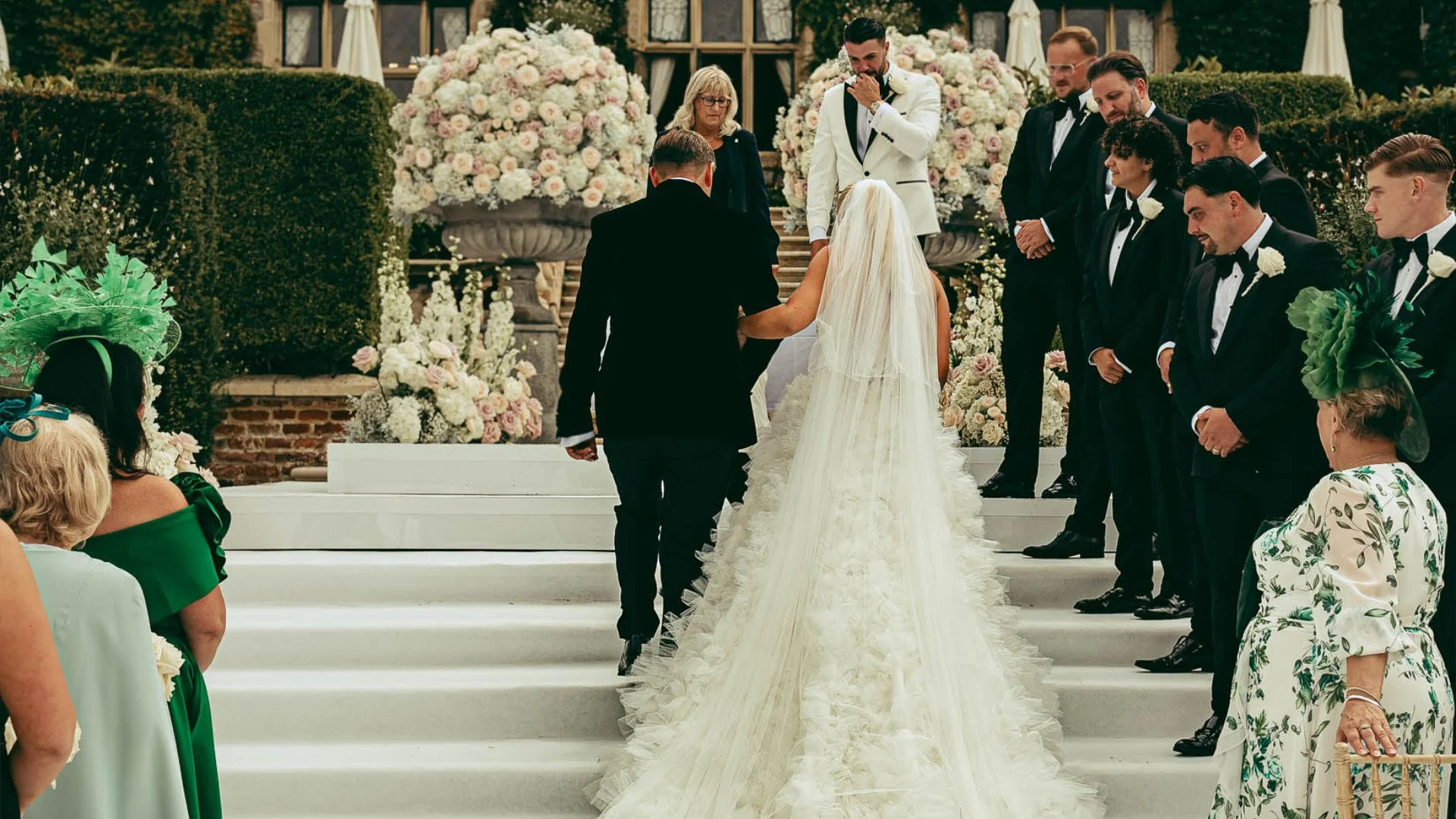 A groom cries as his bride enters their outdoor wedding ceremony and her long luxurious dress flows down white steps, captured by a cinematic wedding videographer.